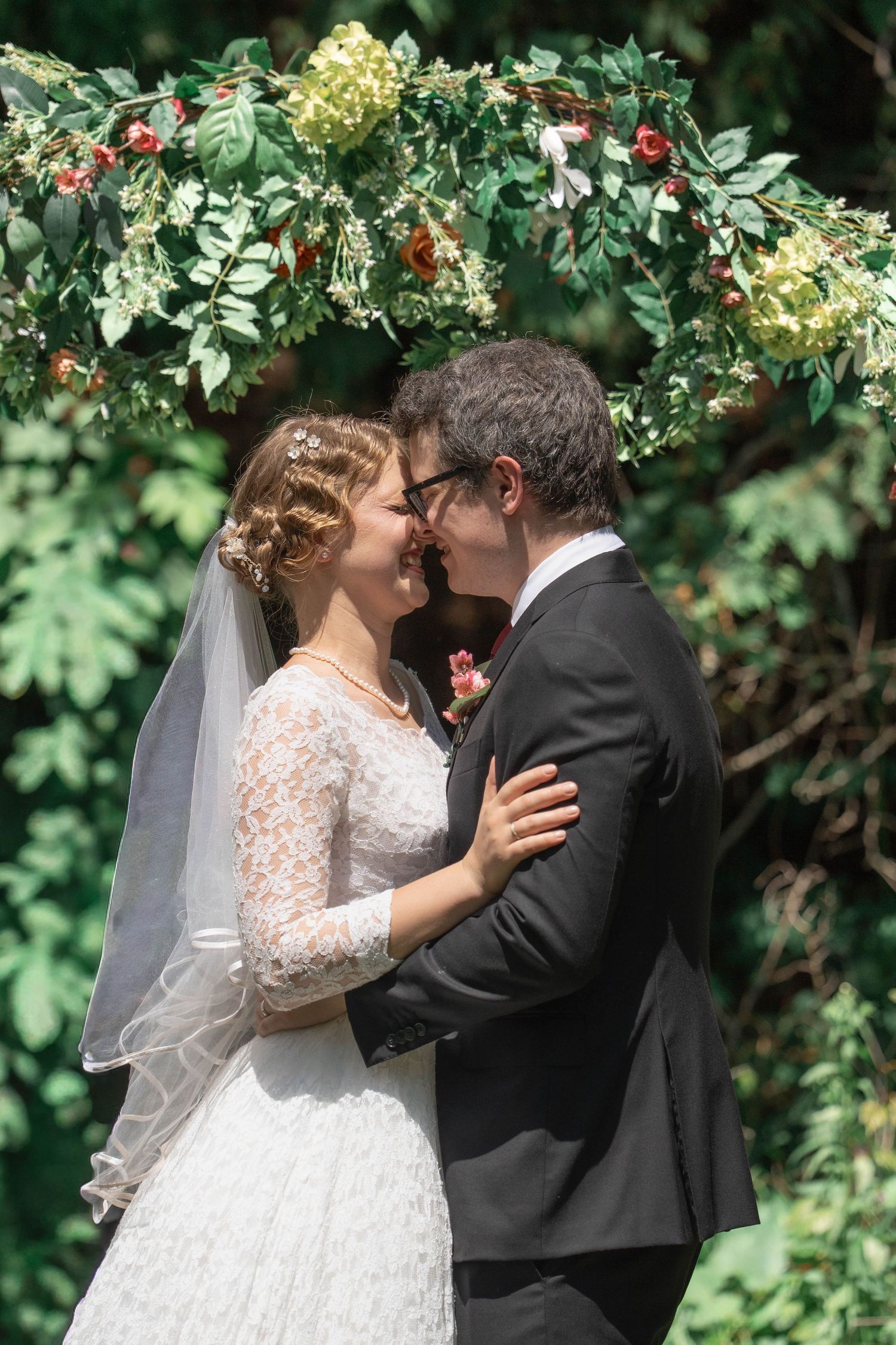 A bride in a lace gown and veil embraces and kisses her groom beneath a floral arch of green leaves and roses.