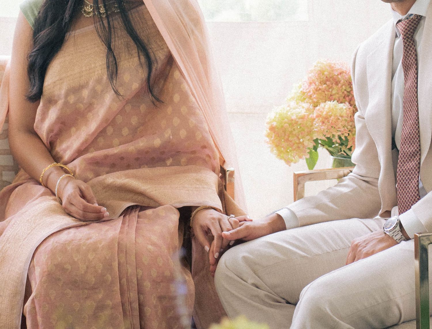 Close-up of a bride in a blush saree and groom in a cream suit sitting together, hands gently touching, beside a peach flower bouquet.