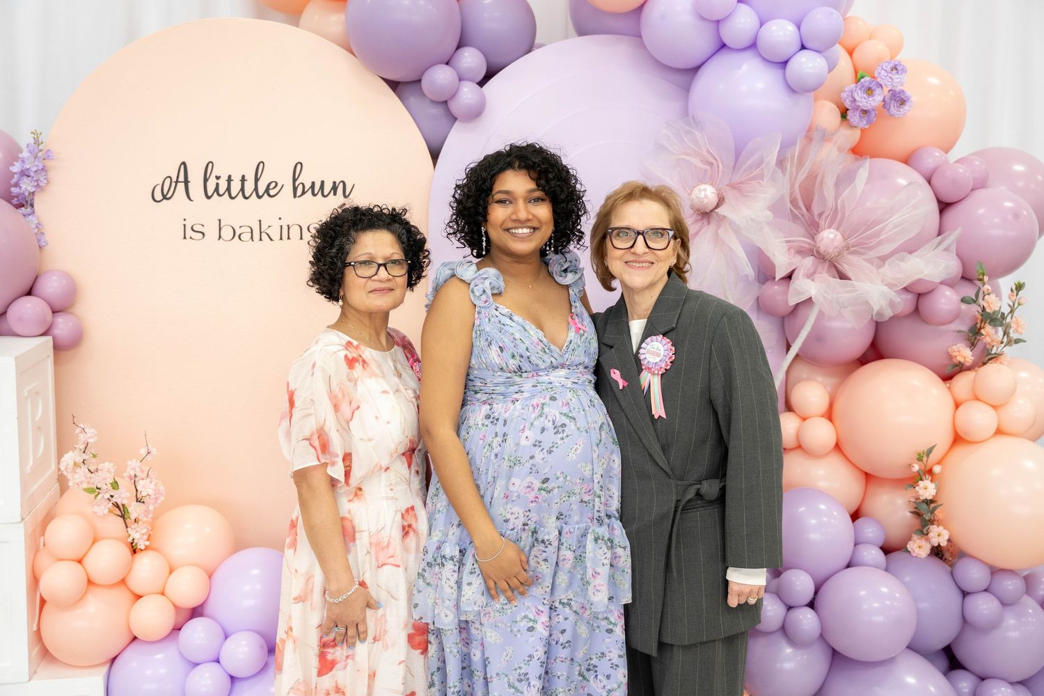 Pregnant woman in a blue floral dress poses between two smiling women in front of a 'A little bun is baking' sign.
