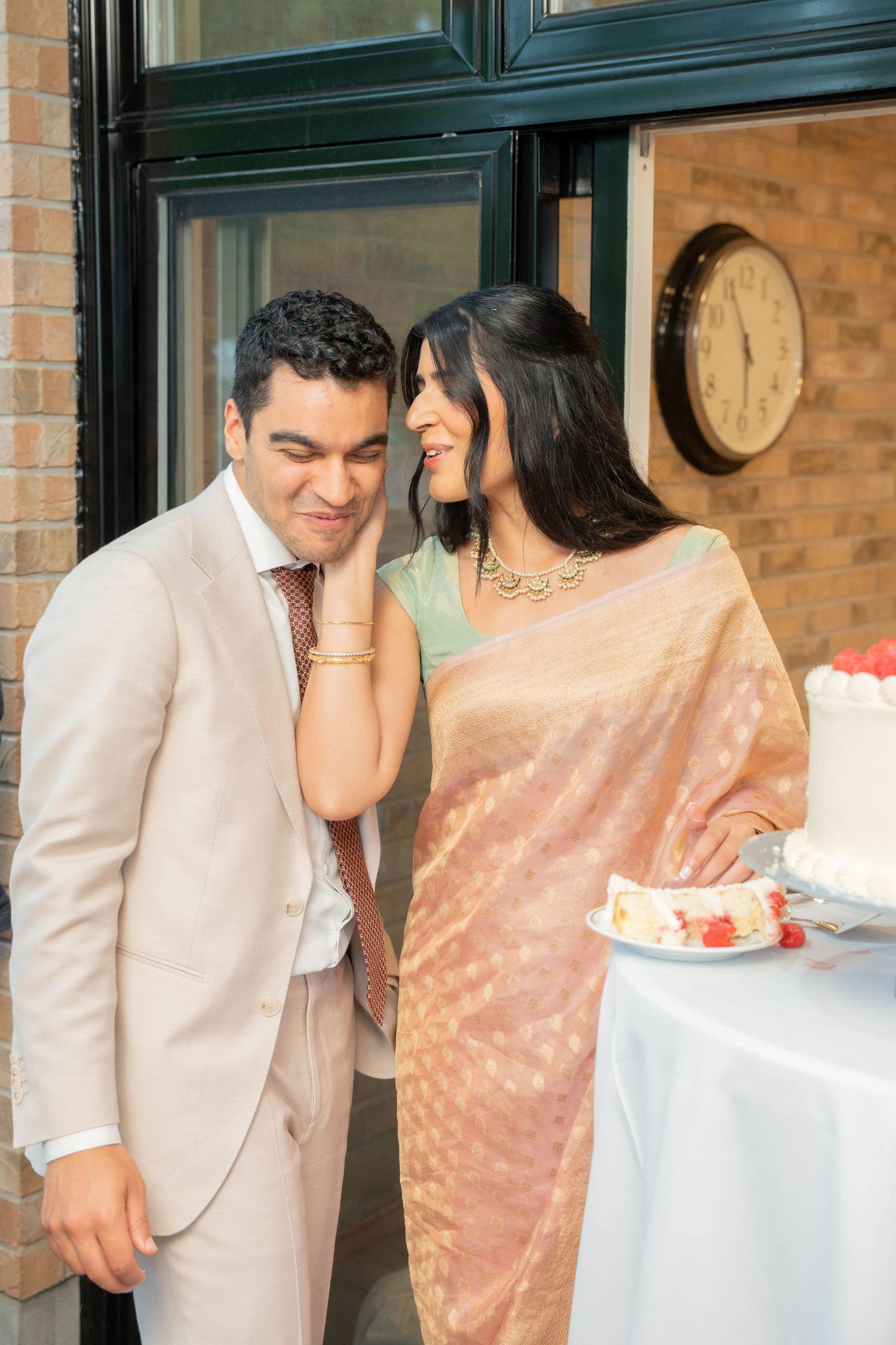 A bride in a peach saree tenderly touches her laughing groom's face beside a wedding cake and a slice on a plate.
