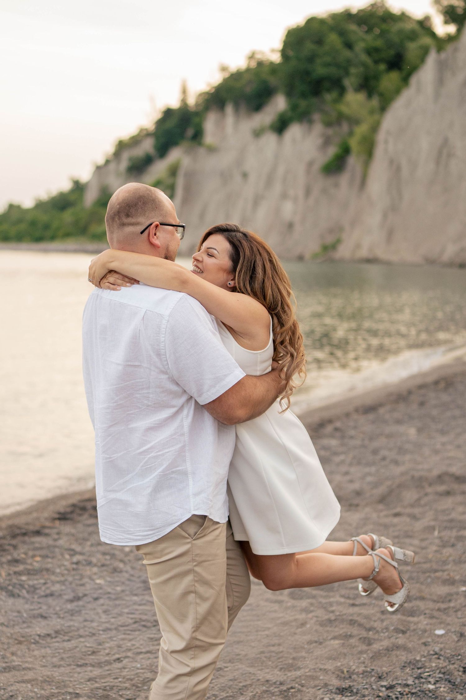 Man in white shirt lifts and hugs his smiling fiancée in a white dress on a beach beside tall coastal cliffs.