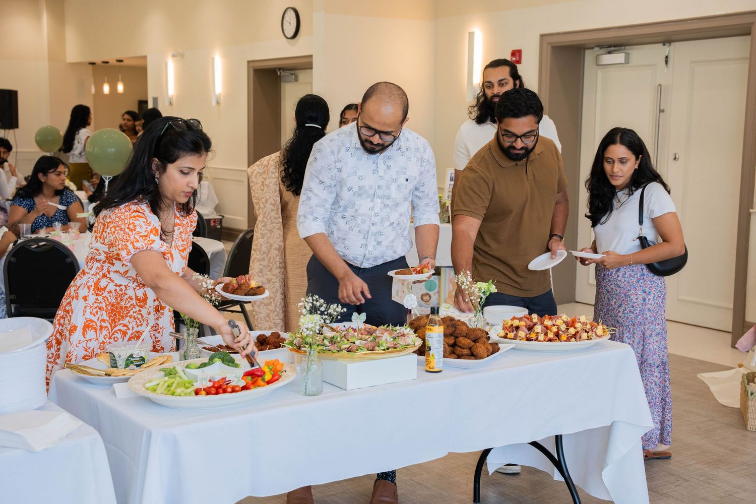Baby shower guests serve themselves from a white-linen buffet of salads, appetizers, and snacks in a bright event room.