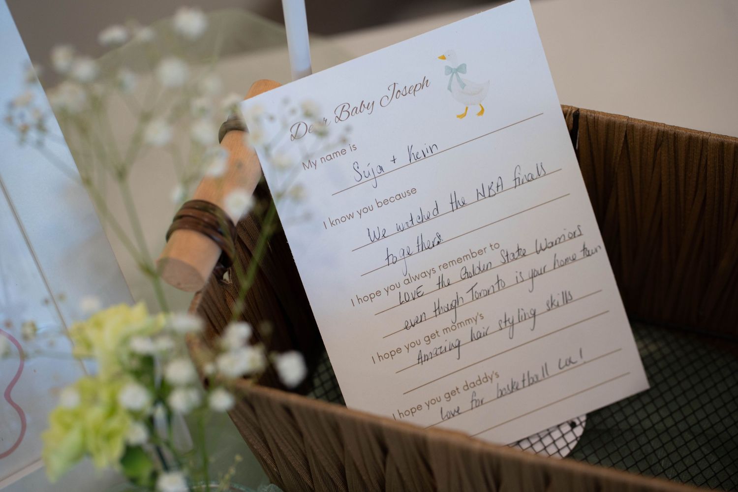 A baby shower advice card with handwritten wishes for the baby rests on a wicker basket beside delicate white and yellow flowers.