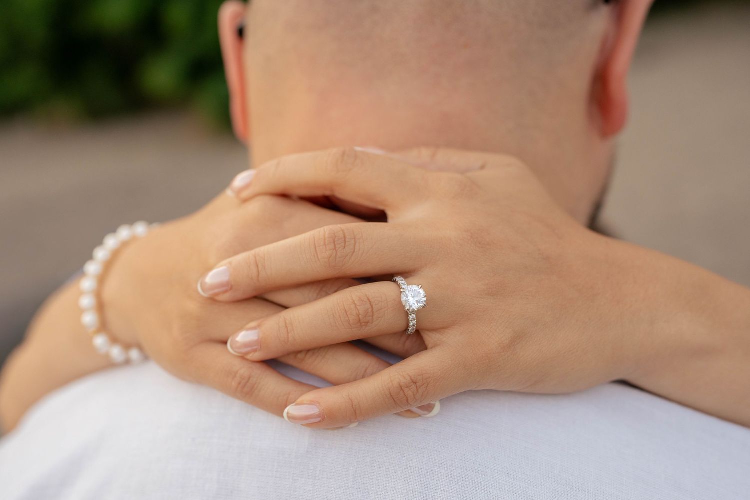 Close-up of a woman's hands wrapped around a man's neck, showing a diamond engagement ring and a pearl bracelet.