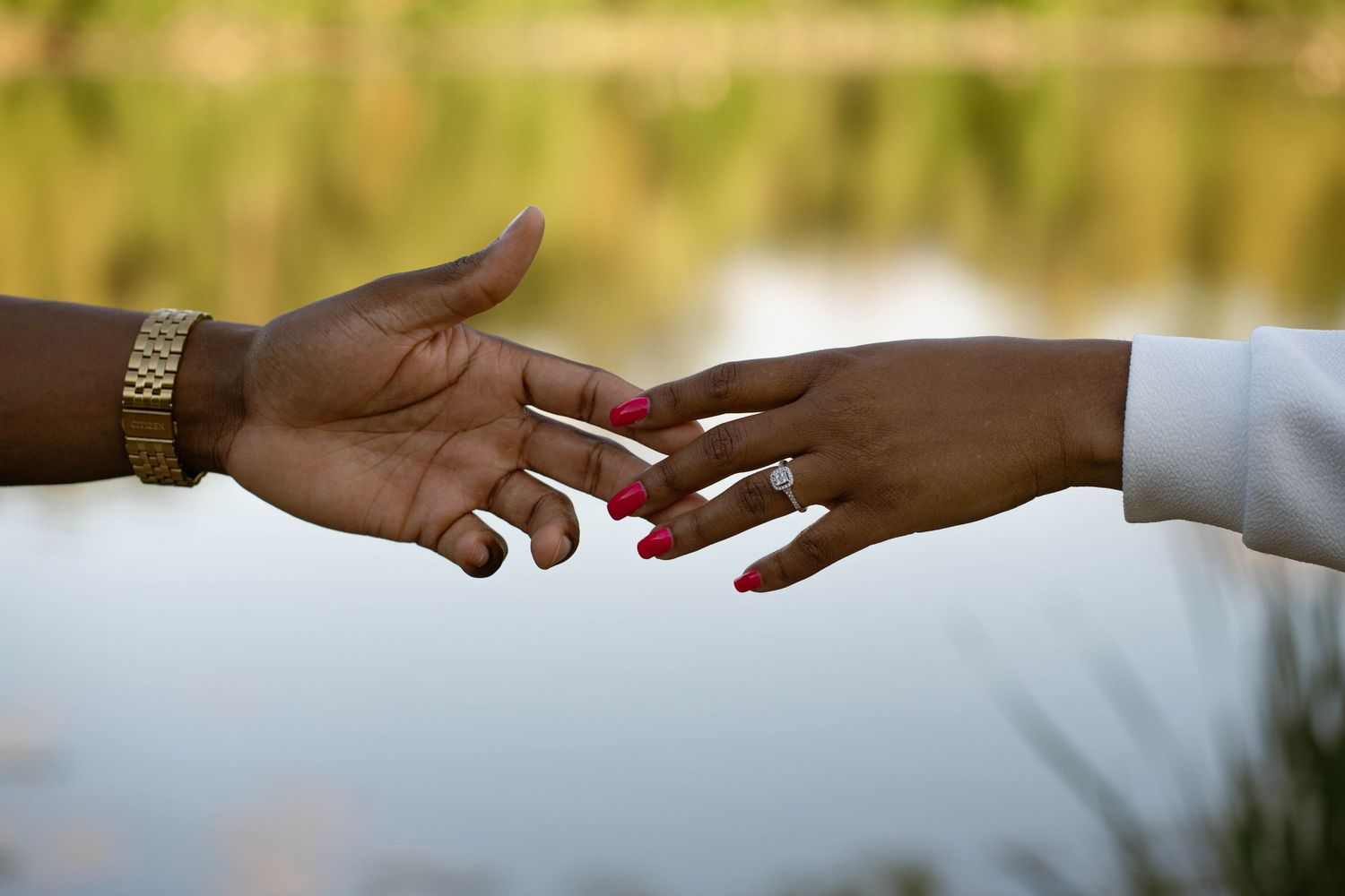 Close-up of two hands reaching toward each other over a calm lake, one wearing a gold watch and the other a ring.