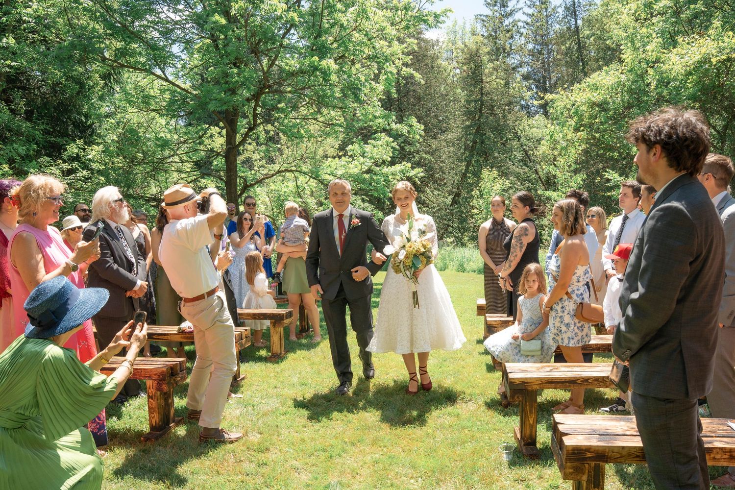 A bride in a white dress carrying a bouquet walks down a grassy aisle between wooden benches and guests at an outdoor wedding.
