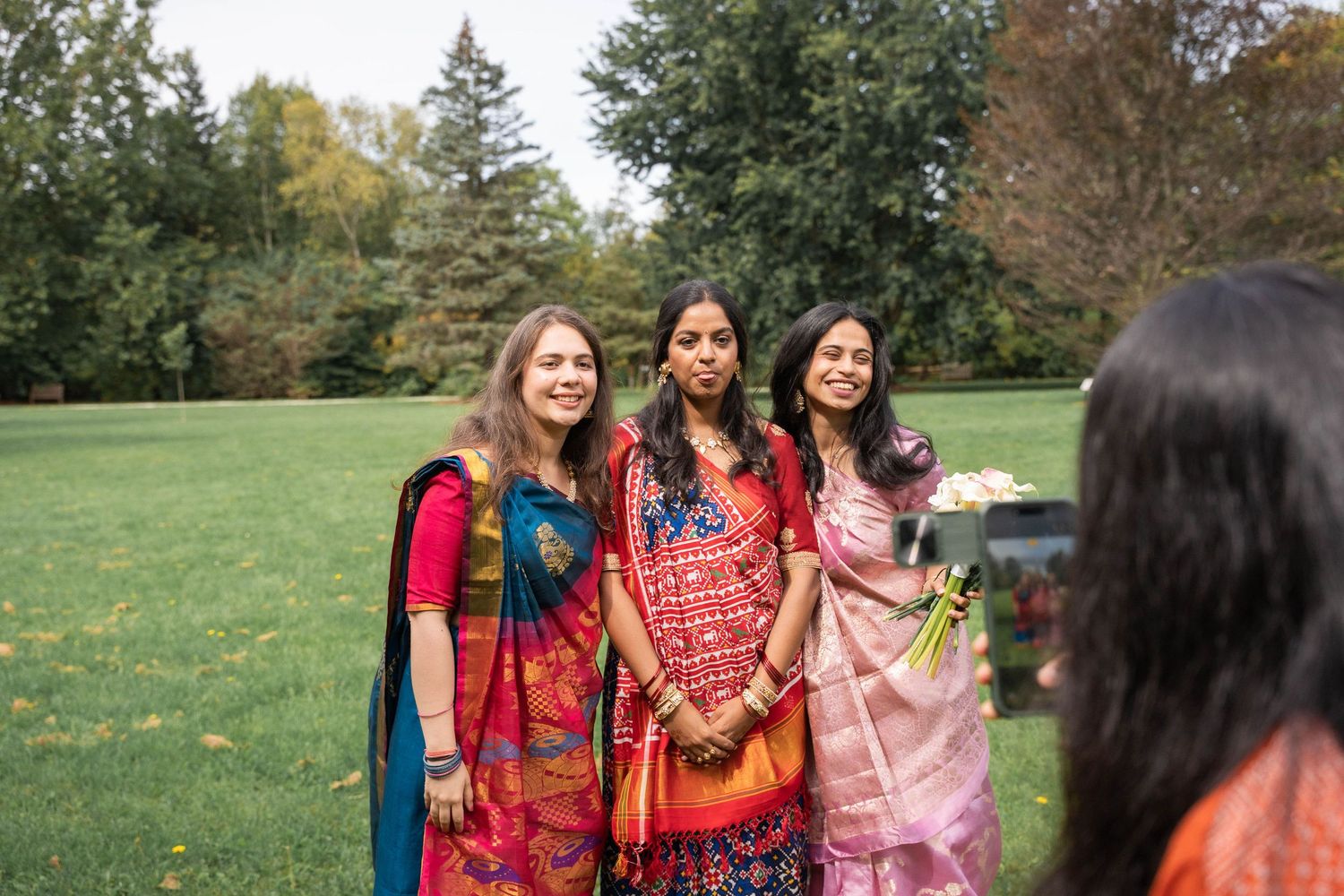 Three women in bright traditional sarees pose for a photo on a sunny lawn while another guest photographs them in the foreground.