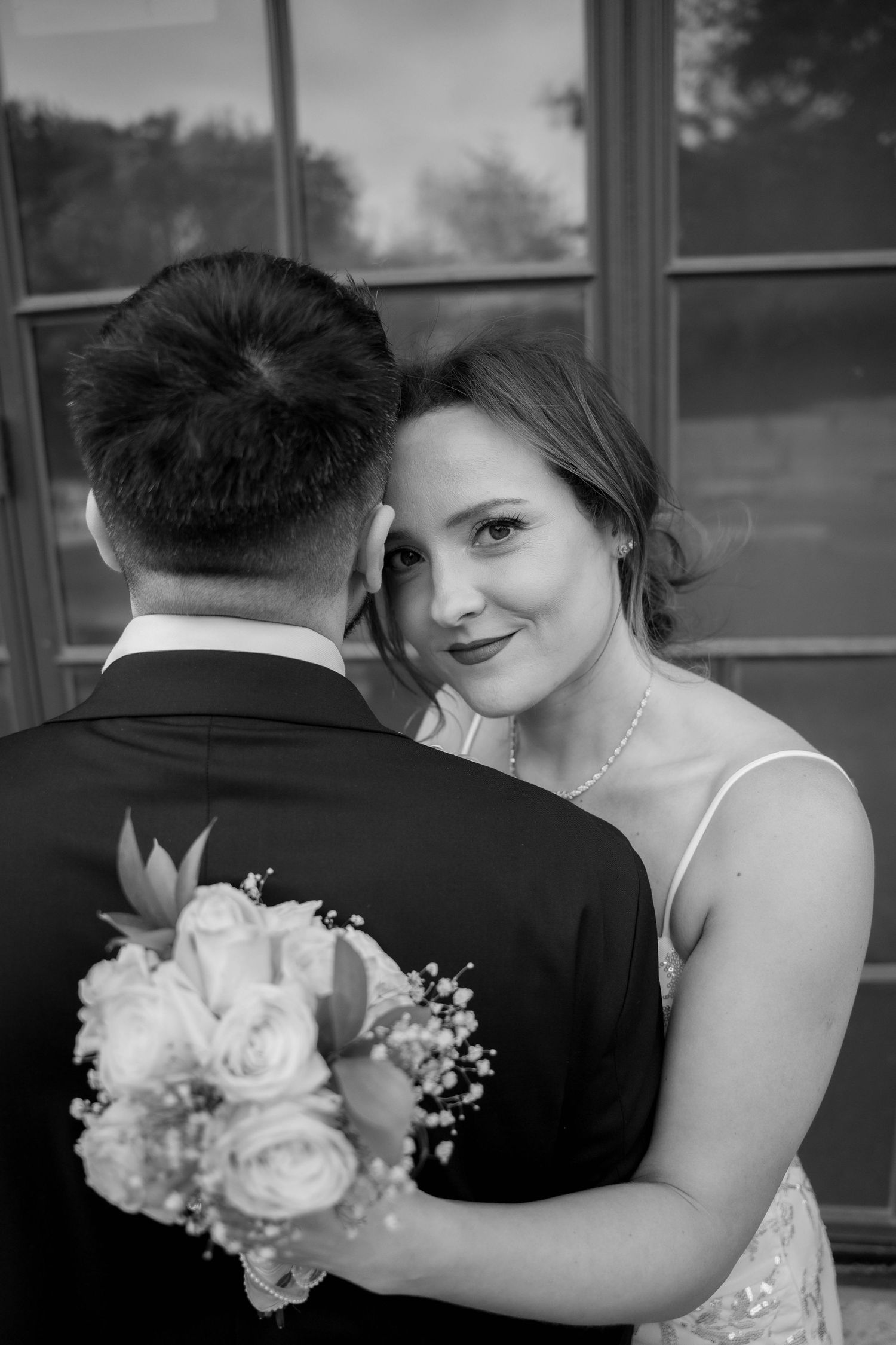 Black and white portrait of a bride leaning on her groom's shoulder, holding a rose and baby's breath bouquet, smiling at the camera.