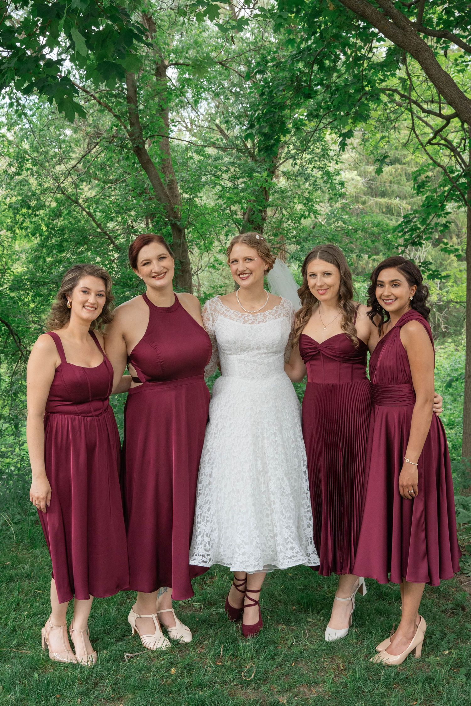 A bride in a white lace dress poses with four bridesmaids in burgundy dresses on a grassy lawn under green trees.