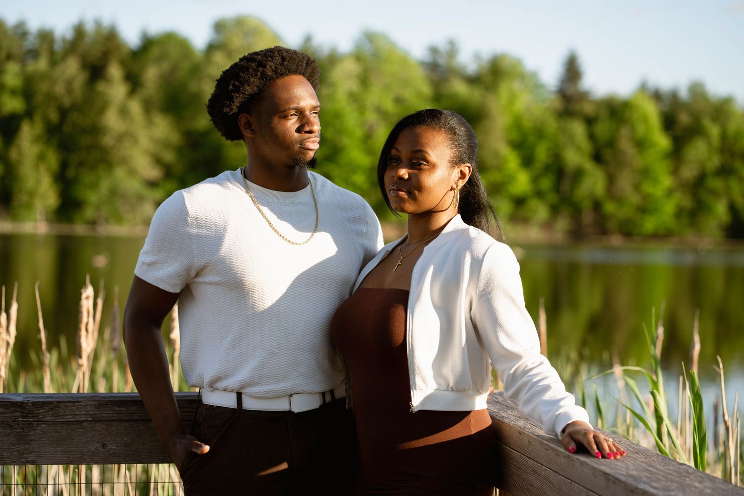 Couple standing together at a wooden railing overlooking a peaceful lake framed by tall green trees at sunset.