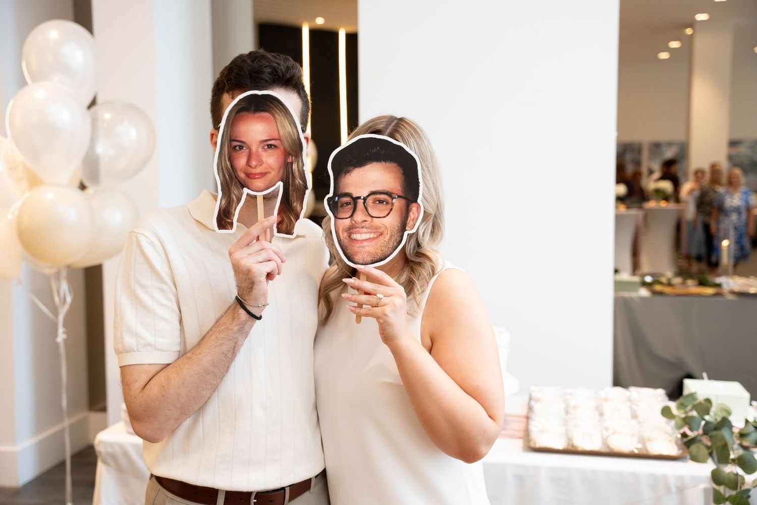 A couple poses cheek to cheek at an engagement celebration, each hiding behind a printed face cutout, with white balloons nearby.
