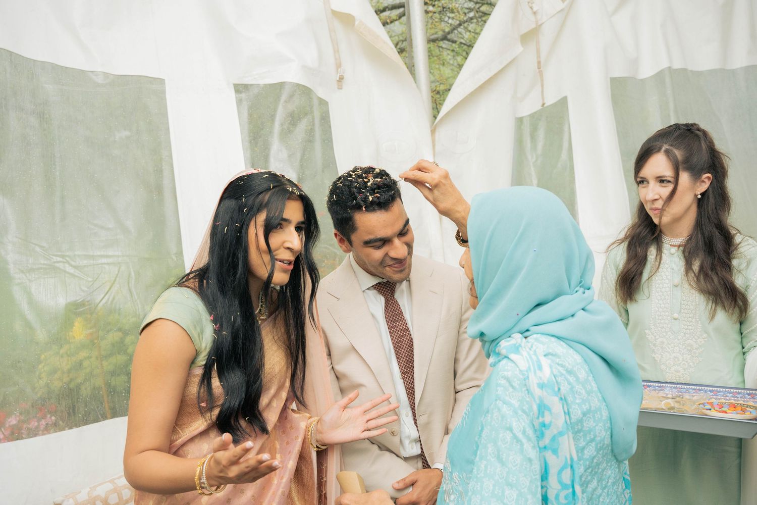 Three women in colorful South Asian attire chat animatedly with a smiling man in a beige suit under a white tent.