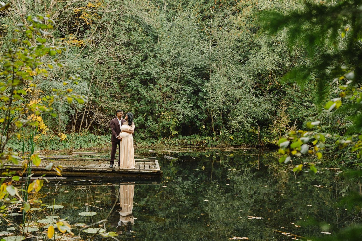 A couple in formal attire embraces on a wooden dock beside a calm pond surrounded by lush green trees.