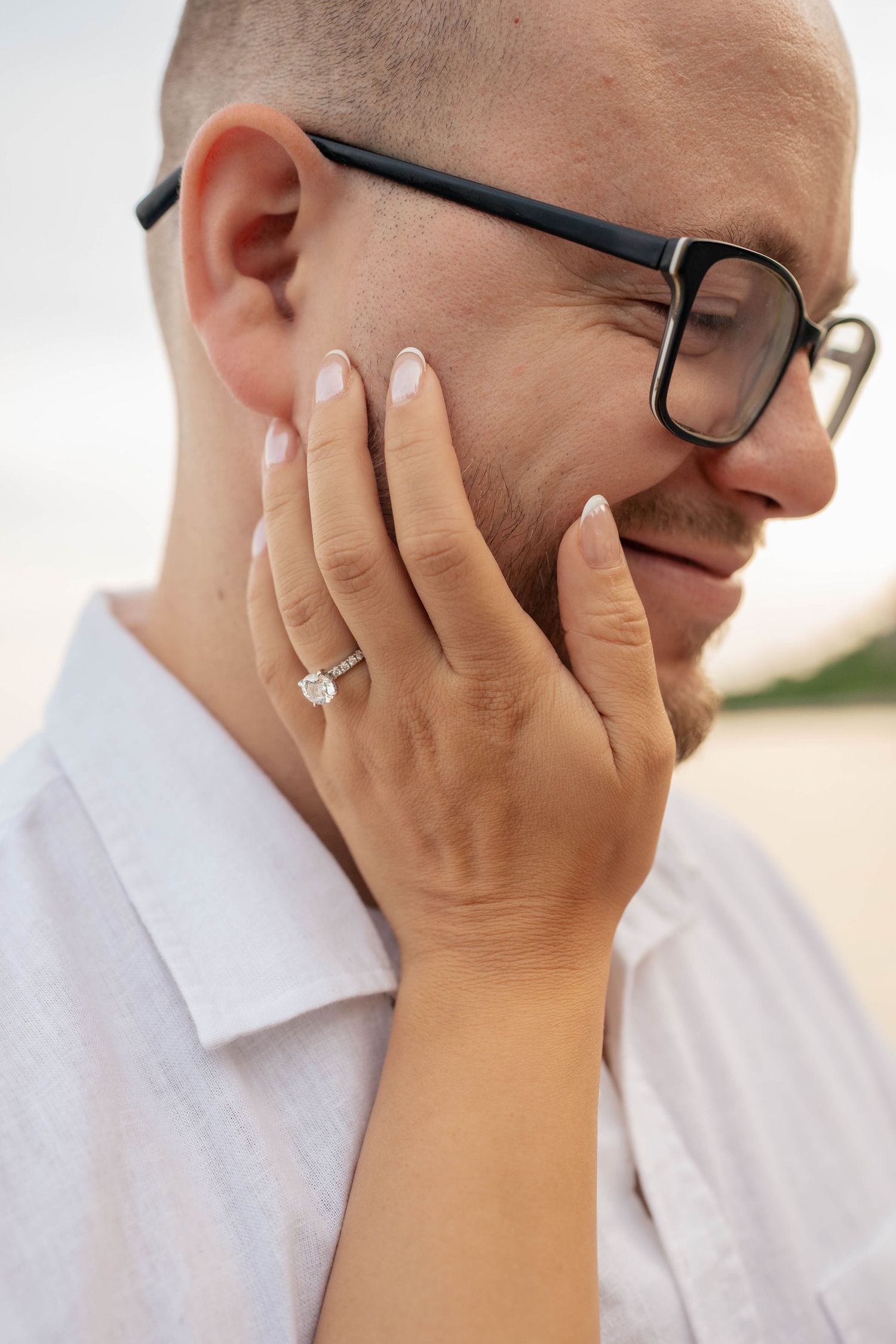 Close-up of a woman's hand with a diamond engagement ring cupping the cheek of a smiling man in glasses.