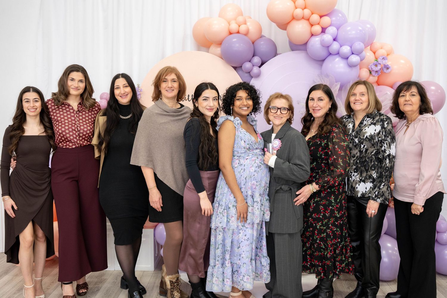 Group of smiling women in elegant outfits poses together in front of peach and lavender balloon decor at a baby shower celebration.