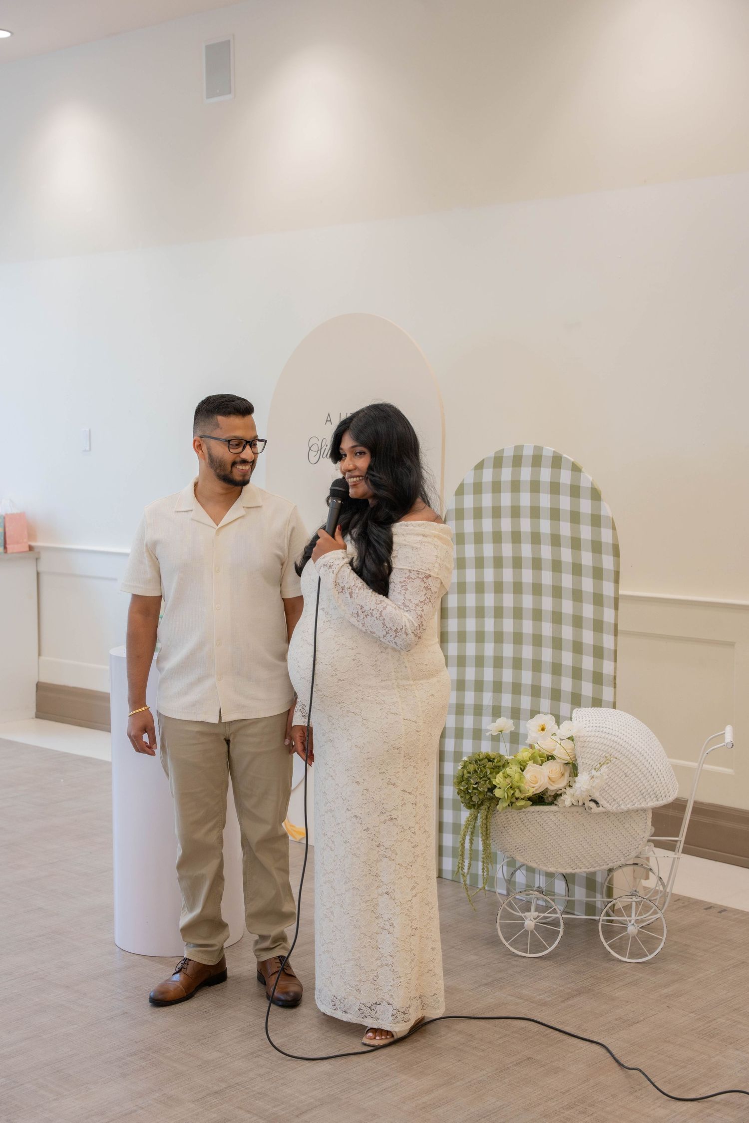 Smiling couple stands beside a green gingham backdrop and floral stroller as the expectant mother speaks into a microphone.