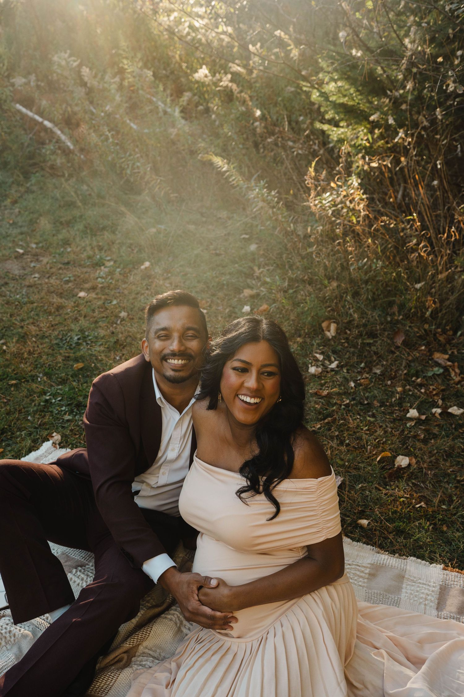 Happy couple laughing together on a picnic blanket in warm sunlight during a maternity photo shoot, with the mother-to-be in a blush dress.