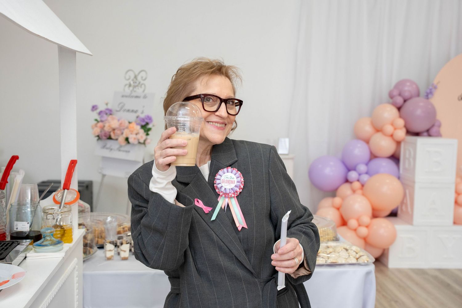 A smiling woman wearing glasses and a pink ribbon badge holds a drink at a baby shower with pastel balloons.