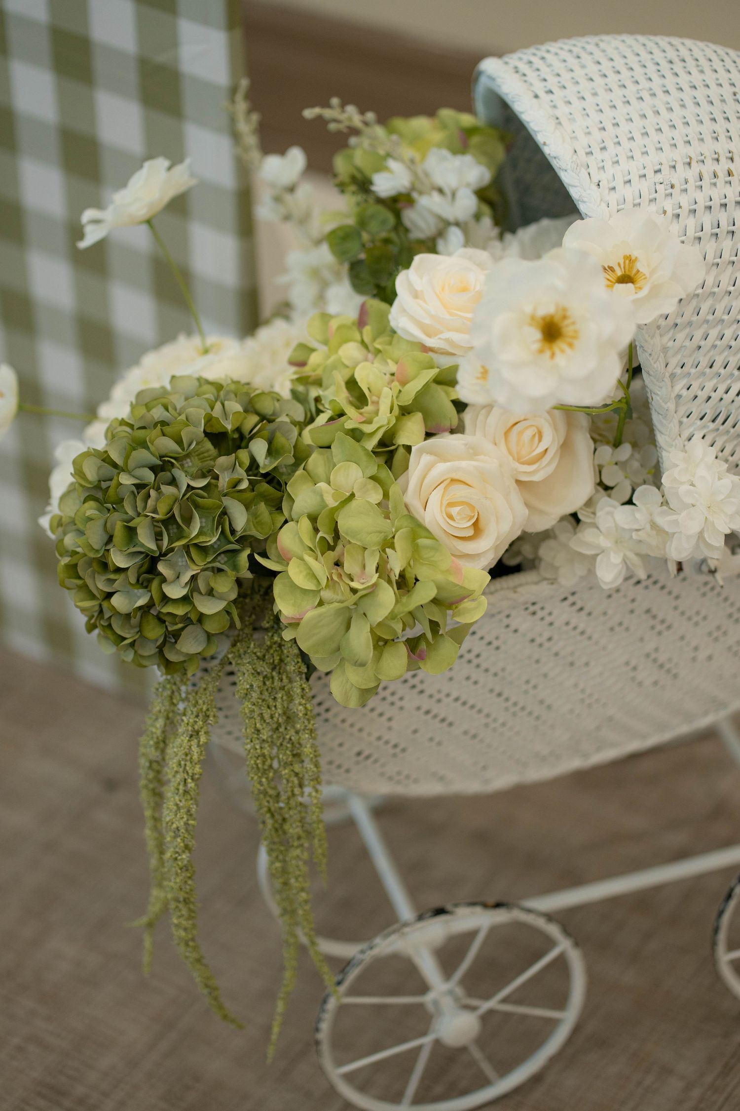 White wicker baby carriage filled with cream roses, green hydrangeas and white blooms beside a green gingham backdrop.