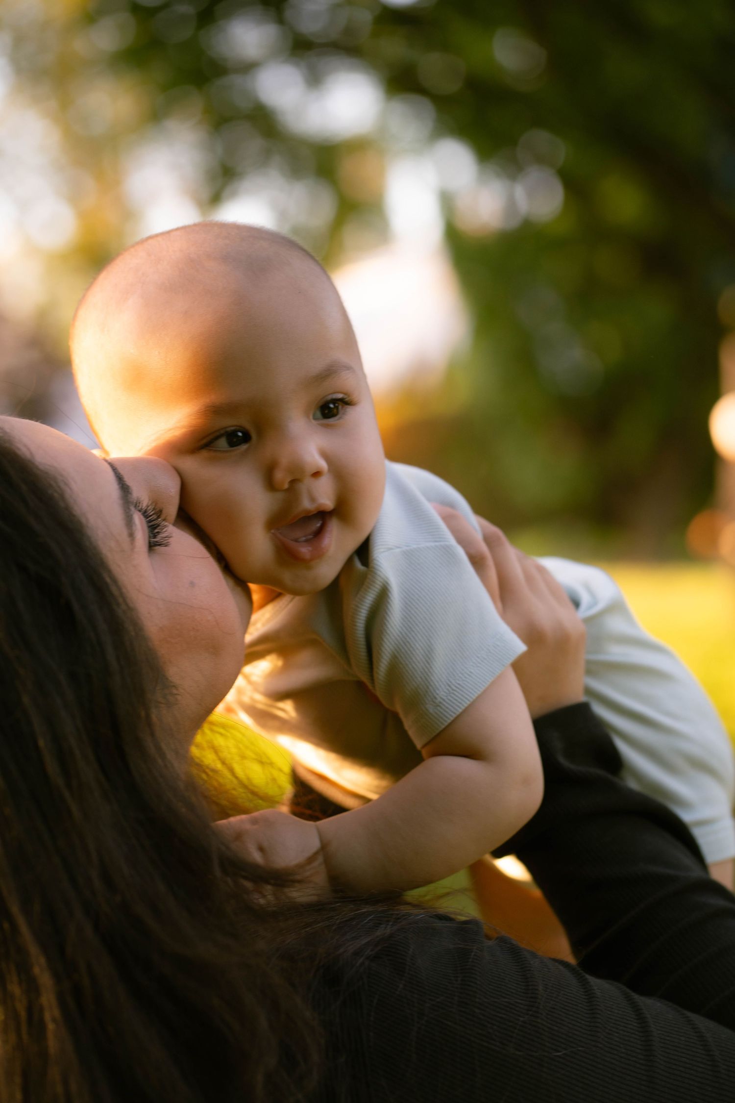 A mother gently kisses her smiling baby's cheek in a park, with soft golden sunlight and blurred trees behind them.