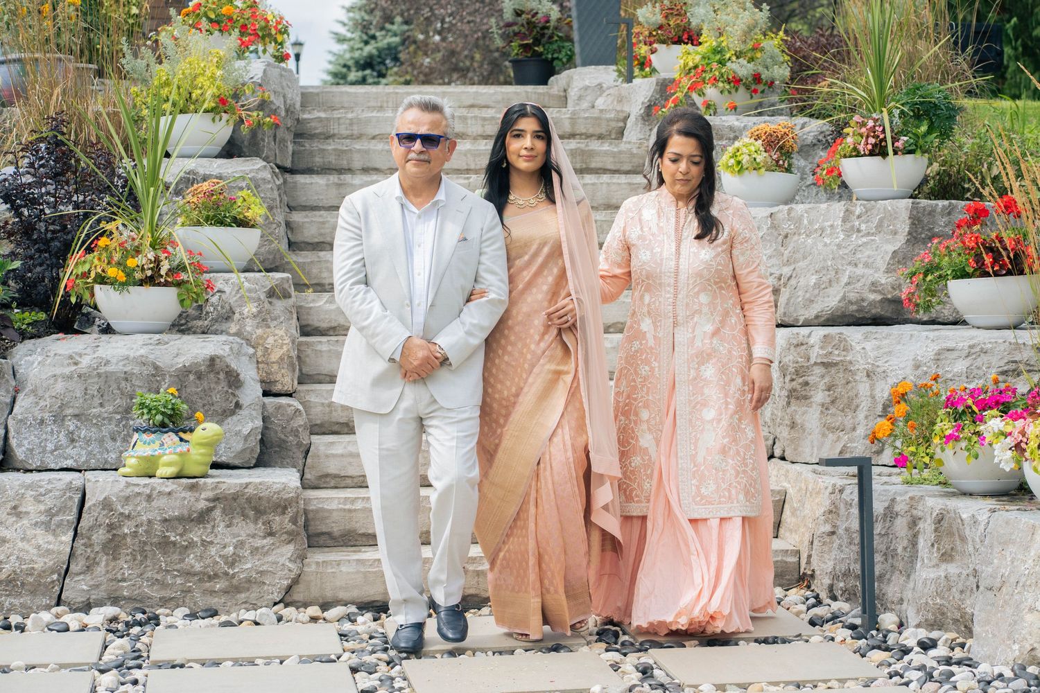 A bride in a peach sari with a veil descends garden steps between her father and a woman, framed by flower-filled stone planters.