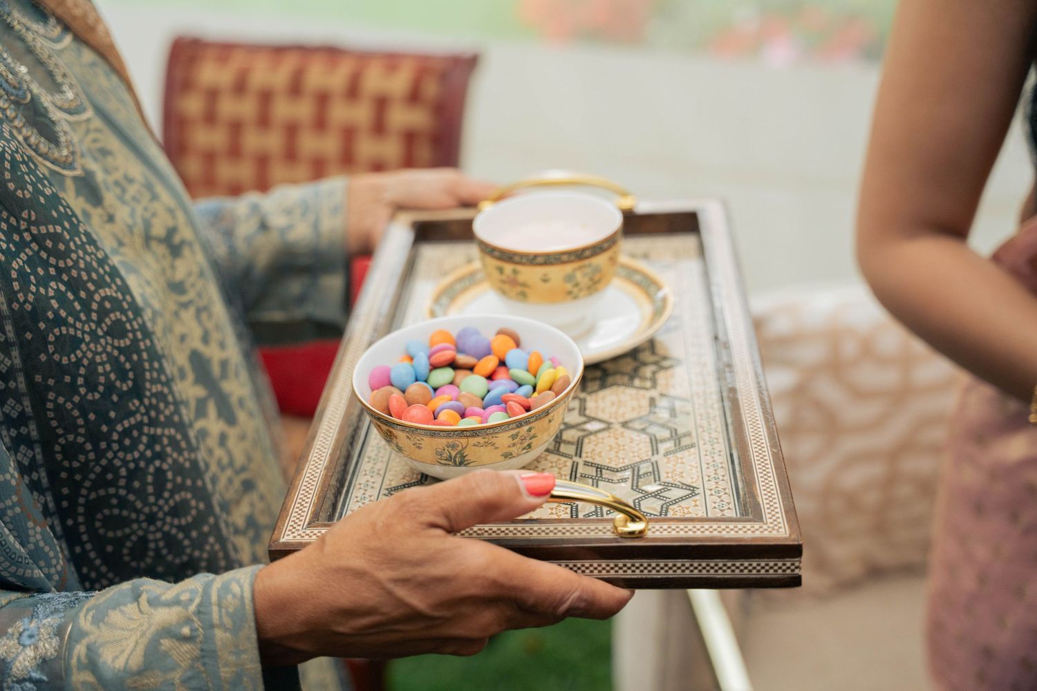 A guest in an embroidered teal tunic holds an ornate tray with a gold-trimmed cup and a bowl of colorful candies at a wedding celebration.