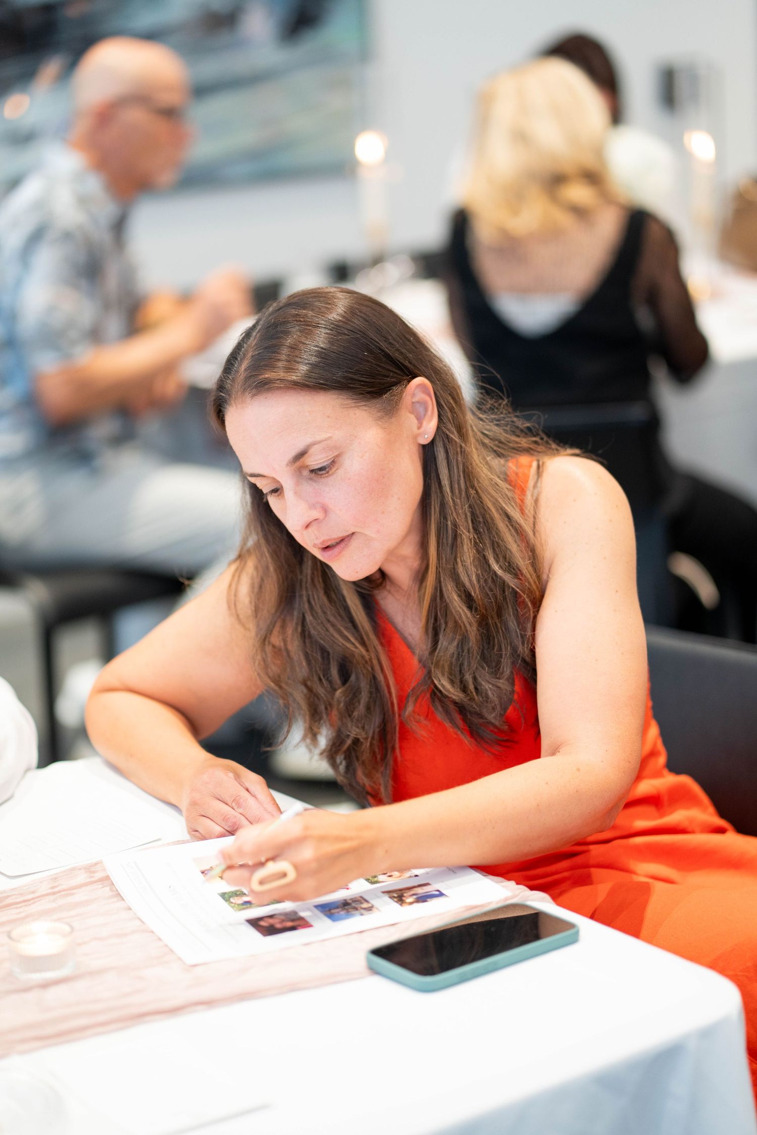A woman in an orange dress marks a photo matching game sheet at a candlelit table during an engagement celebration.