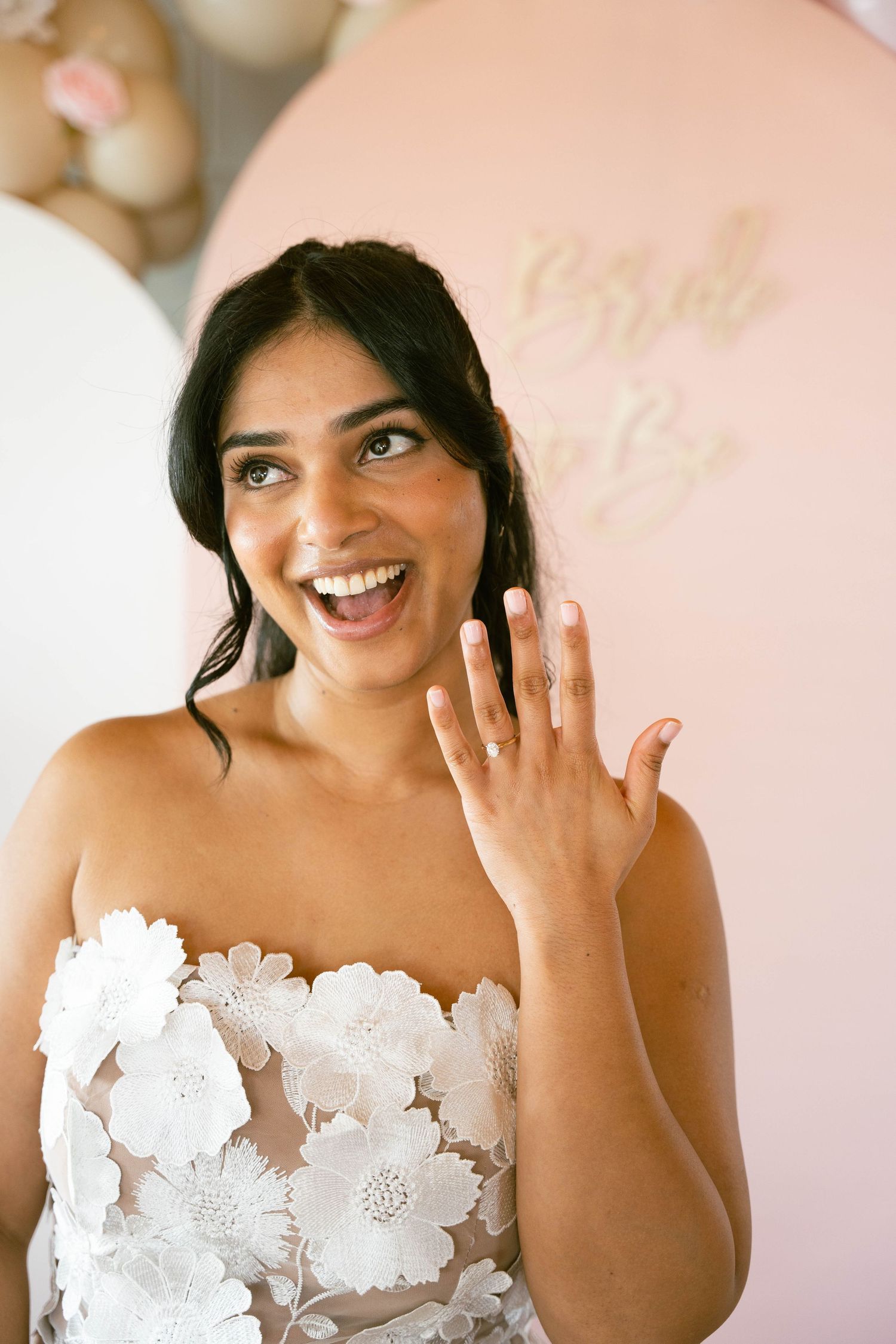 A laughing bride-to-be raises her hand to display her ring at a pink and white bridal shower with a Bride to Be sign.