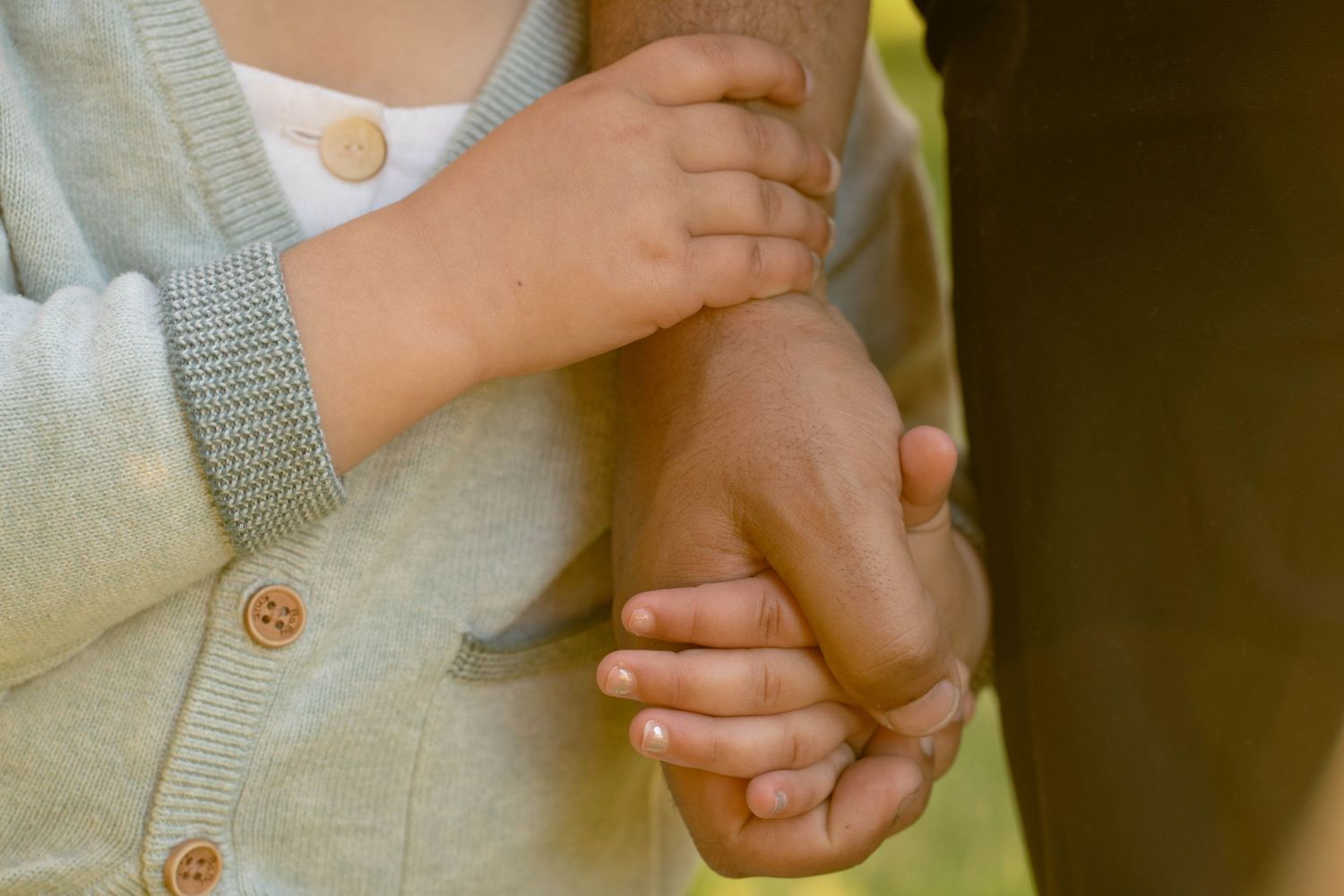 Close-up of a child's small hand resting on a parent's hand, with a gray knit cardigan and wooden buttons visible.