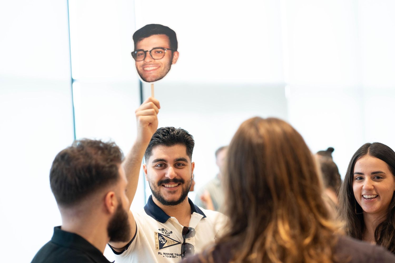 Guest in a white polo holds up a printed cutout of a man's face on a stick as other guests look on at a party.