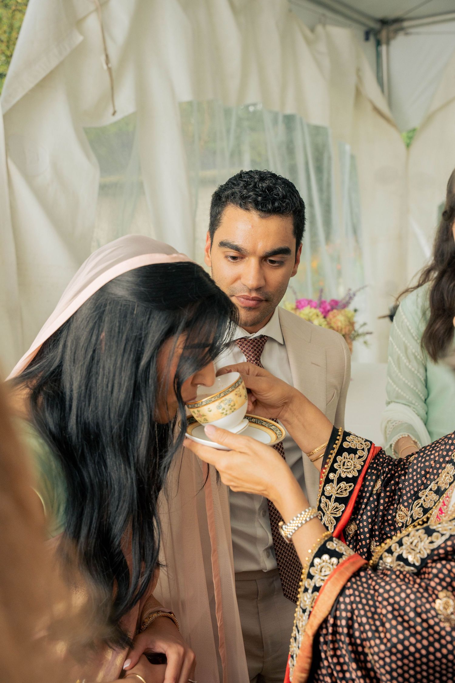 A woman in a pink headscarf sips from an ornate gold-rimmed teacup as a smiling man in a tan suit watches at a traditional wedding ceremony.