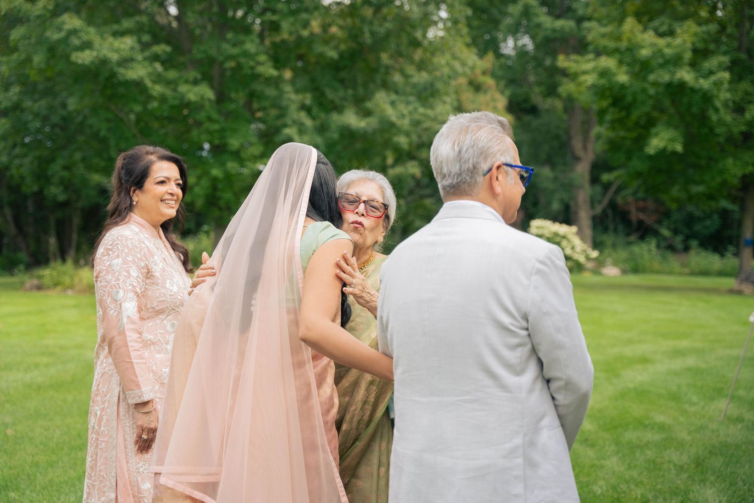 A bride in a peach sari and veil is embraced by family members on a green lawn during an outdoor wedding celebration.