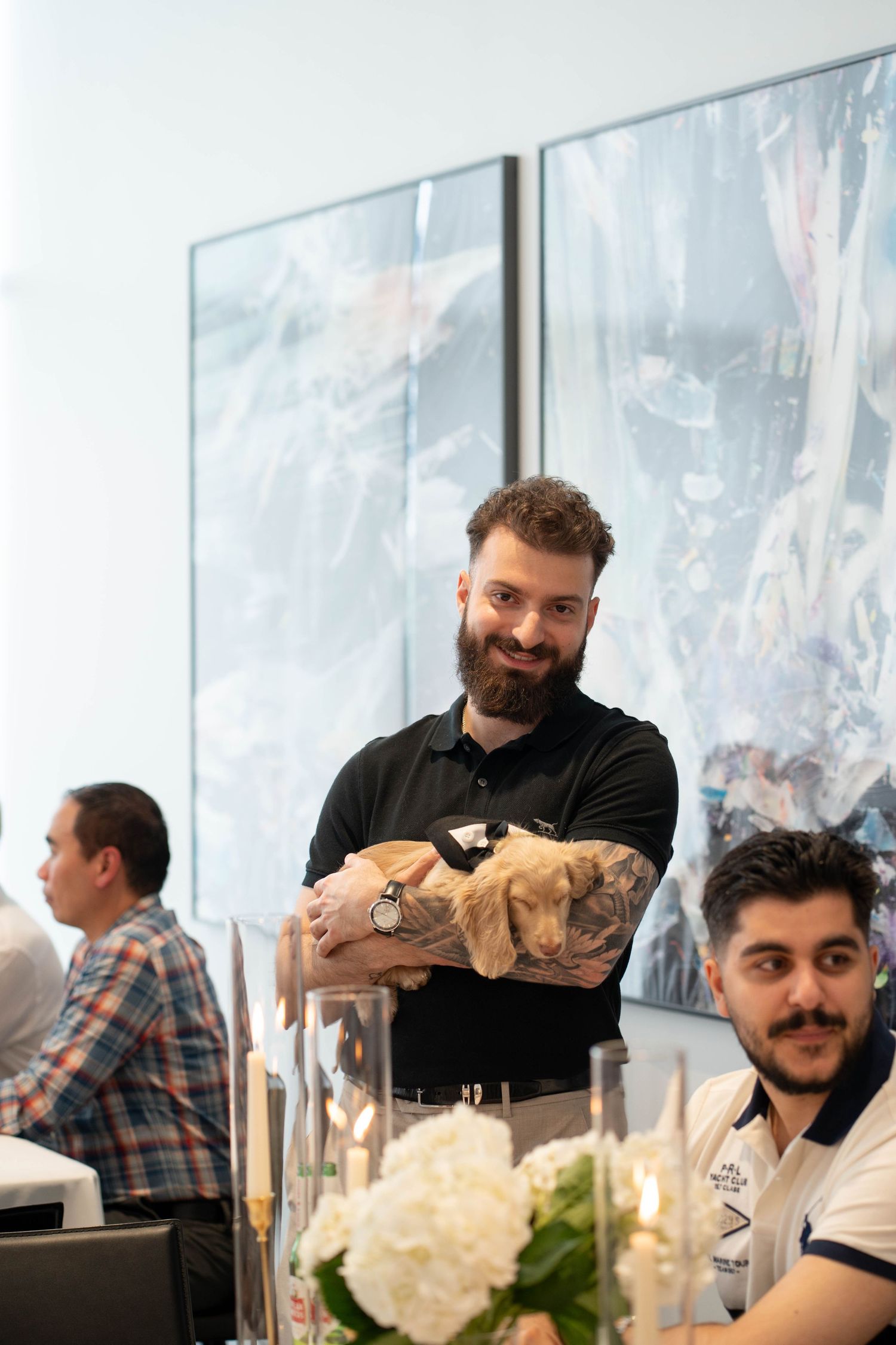 Bearded man in a black polo with tattooed forearms holds a small puppy while standing behind a table with white flowers.