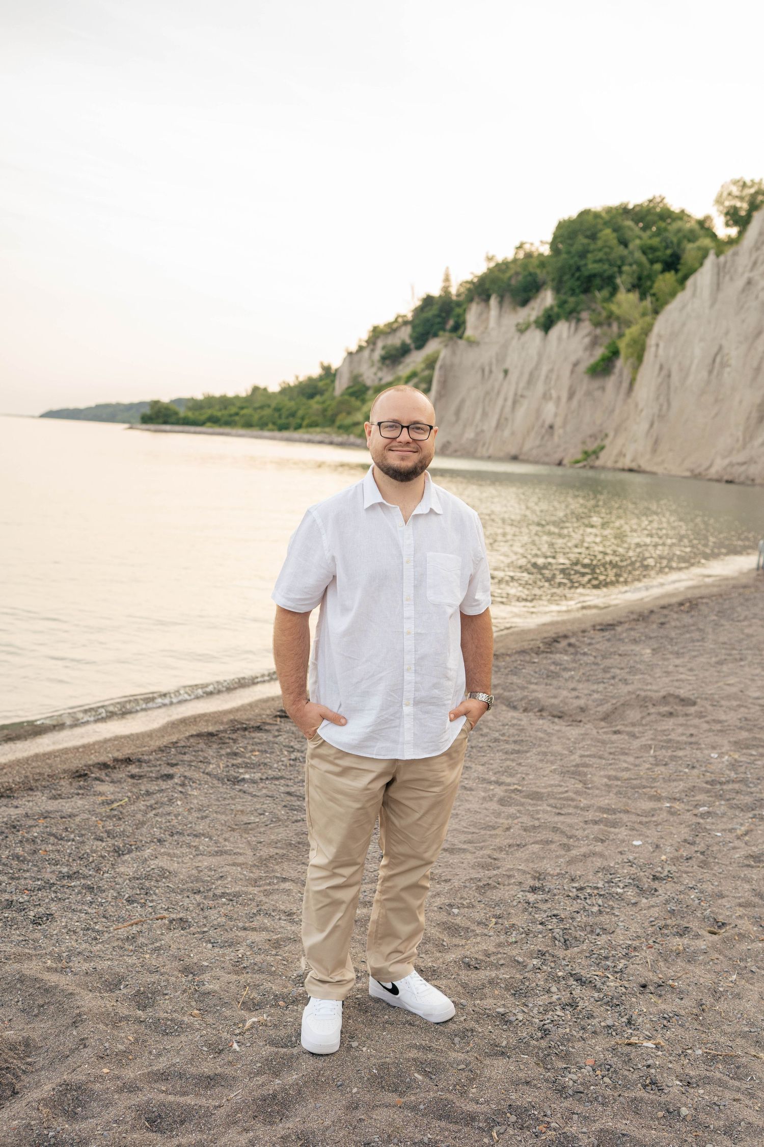 Bearded man in glasses, white linen shirt and khaki pants stands with hands in pockets on a beach below bluffs.