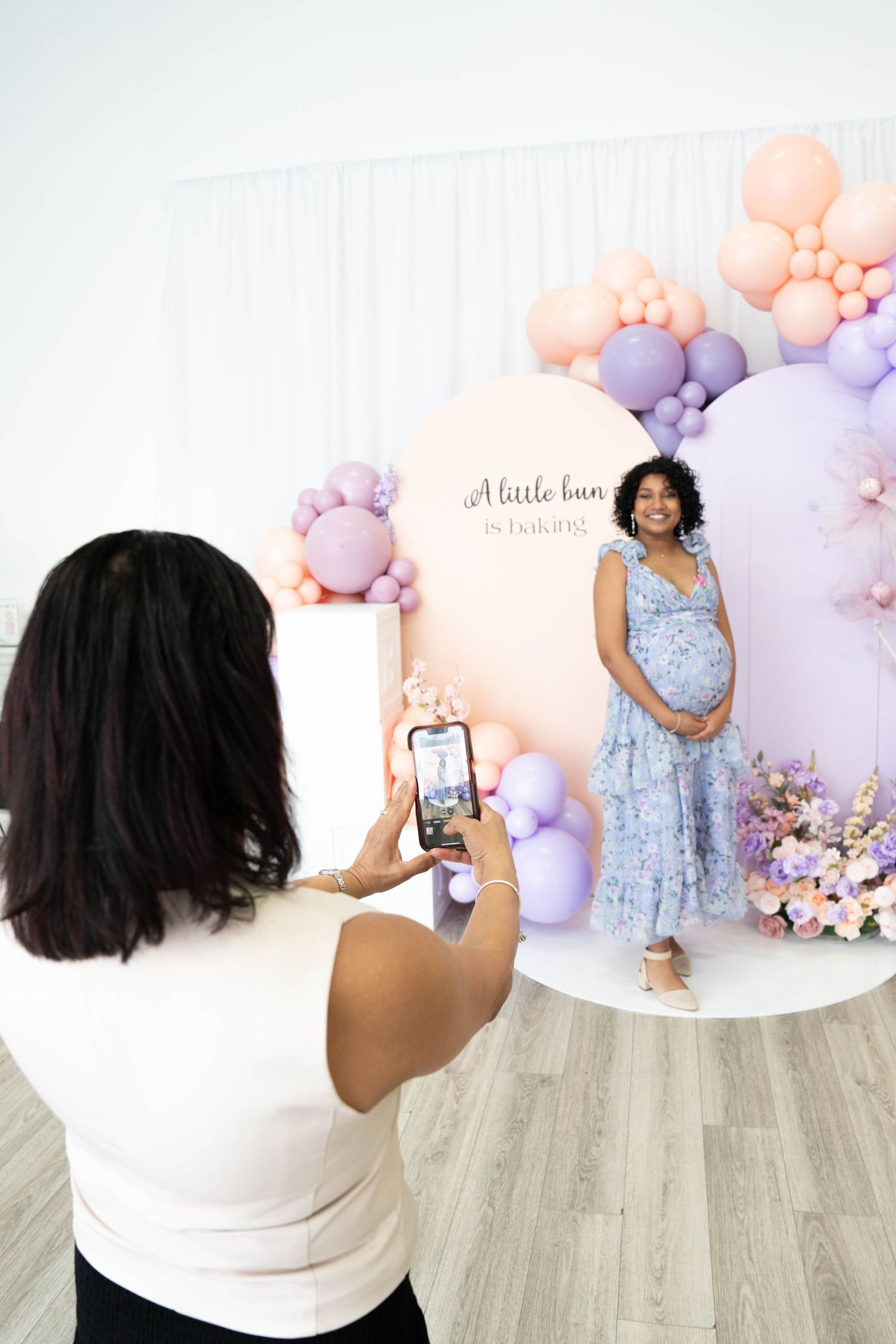 Woman uses her phone to photograph an expecting mother posing in a floral dress before pastel balloons at a bun-in-the-oven baby shower.