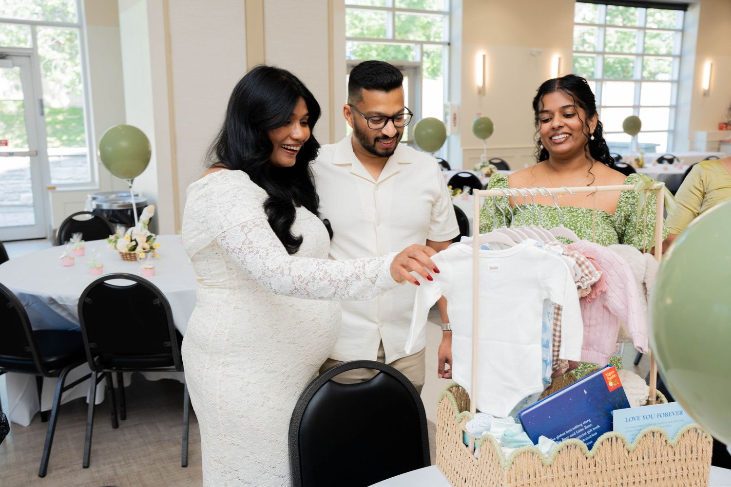 Expectant mother in a white lace dress and her partner look through a gift basket of baby clothes with a smiling guest nearby.