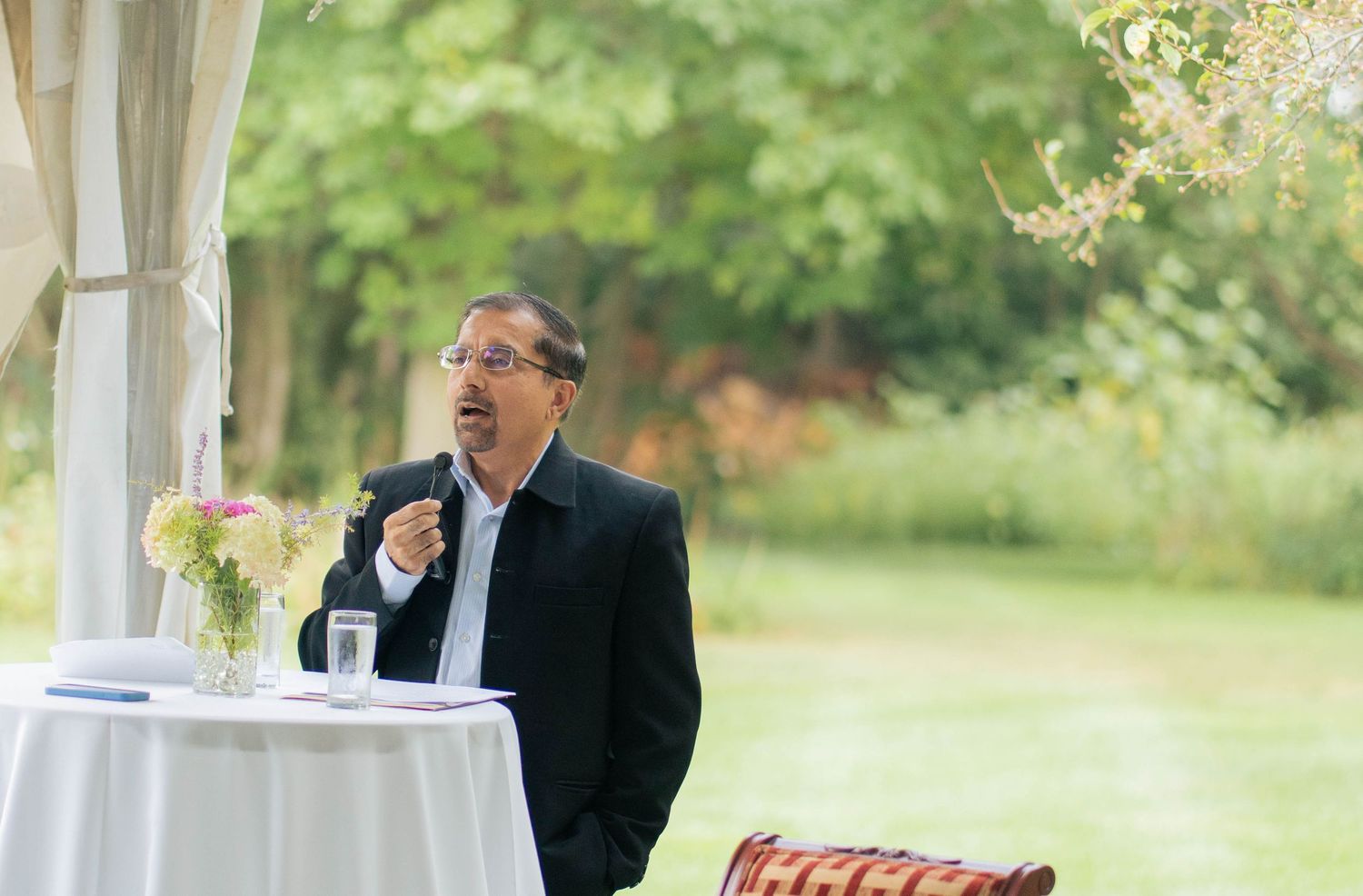 A man in glasses and a black blazer speaks at a draped table with a flower vase during an outdoor wedding ceremony.