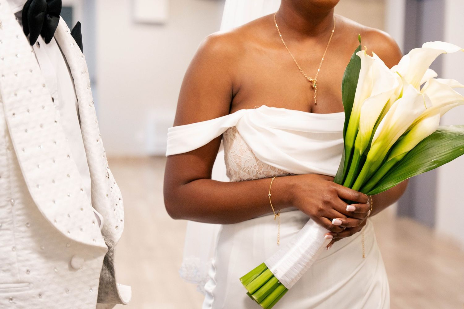 Bride in an off-shoulder white gown holds a bouquet of white calla lilies beside a groom in an embellished ivory jacket.