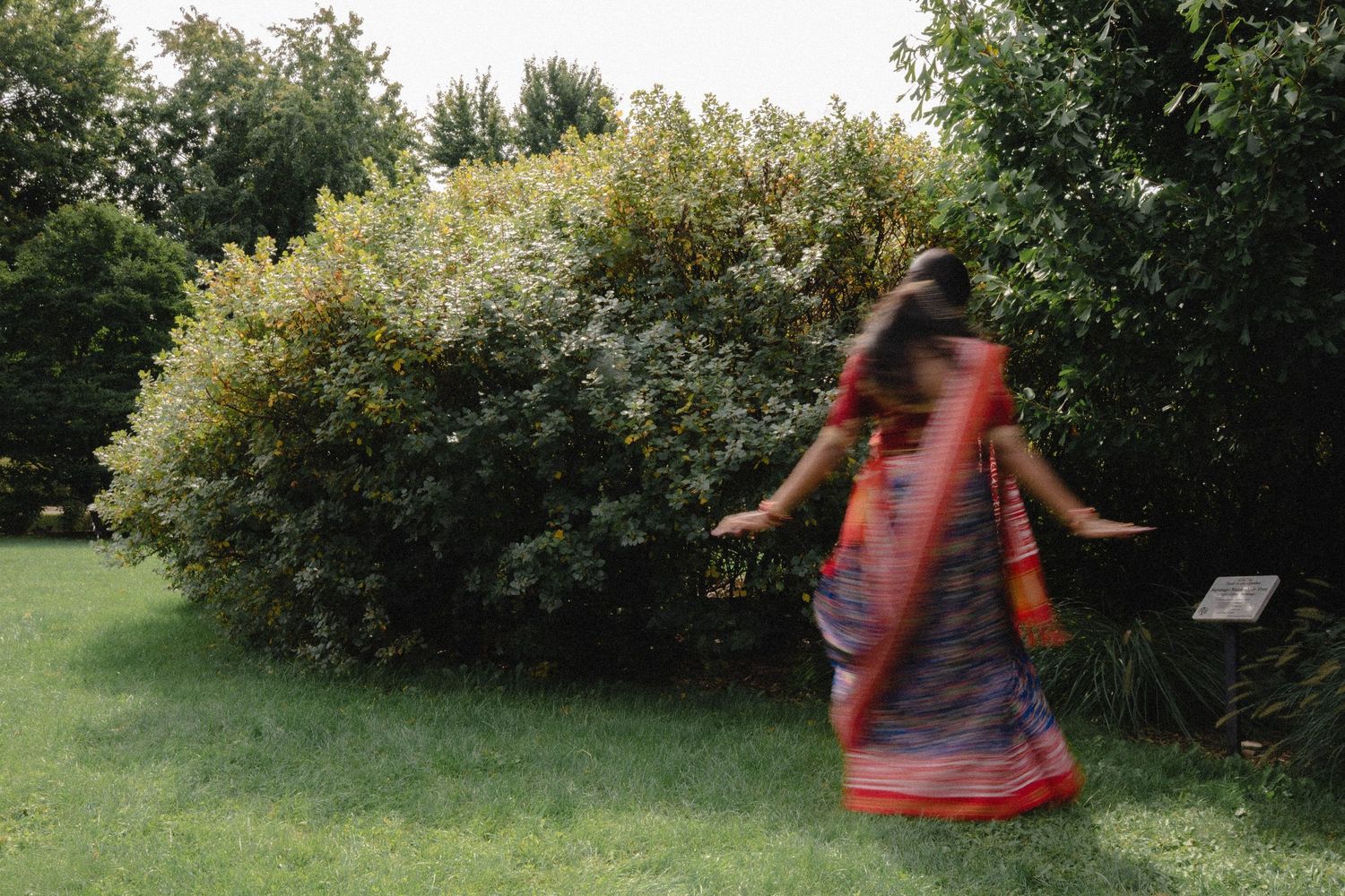 A woman in a flowing saree spins on a green lawn in front of lush shrubs, creating a joyful motion blur.
