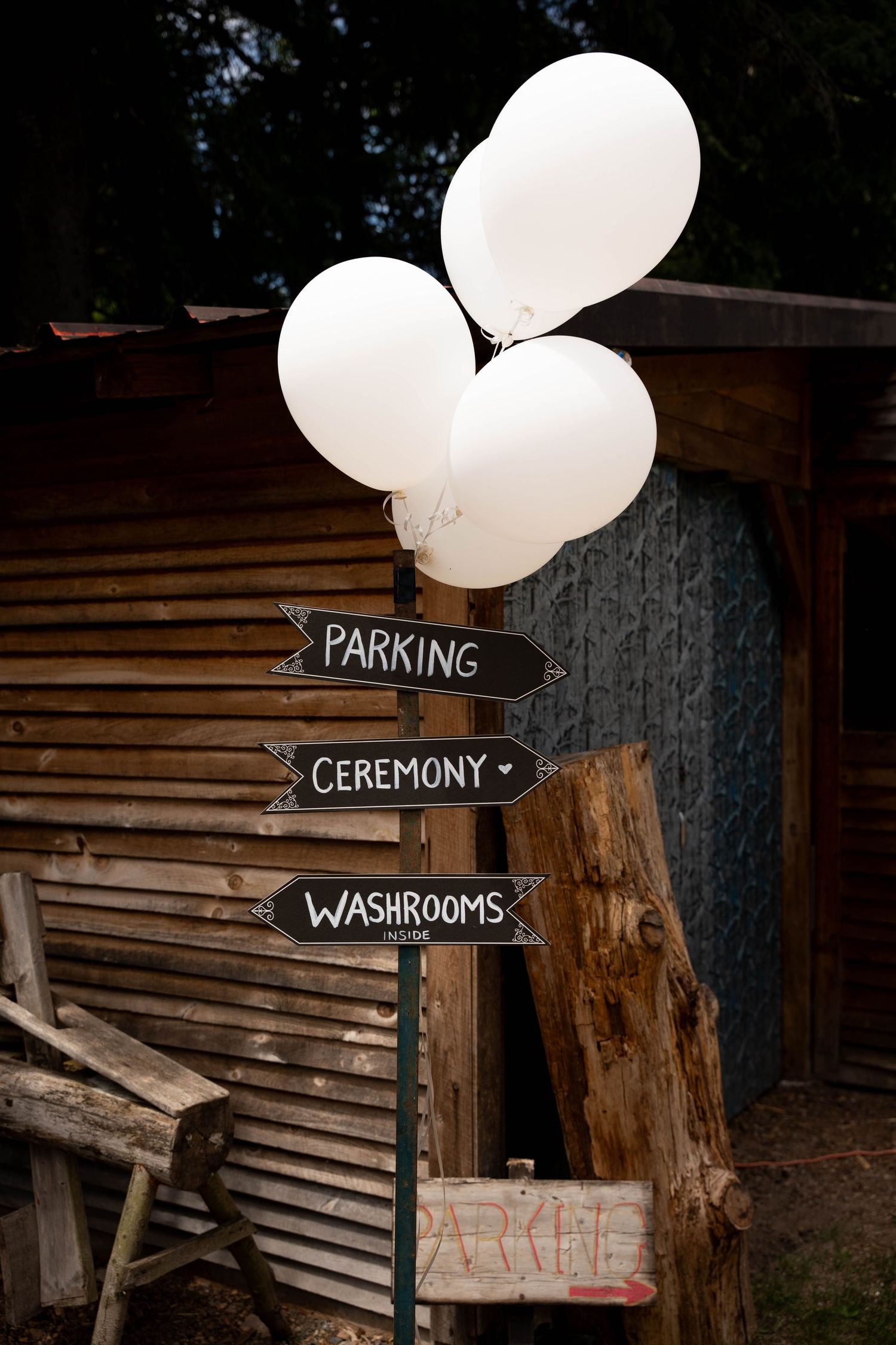 A wooden signpost with chalkboard arrows for parking, ceremony and washrooms is topped with white balloons.