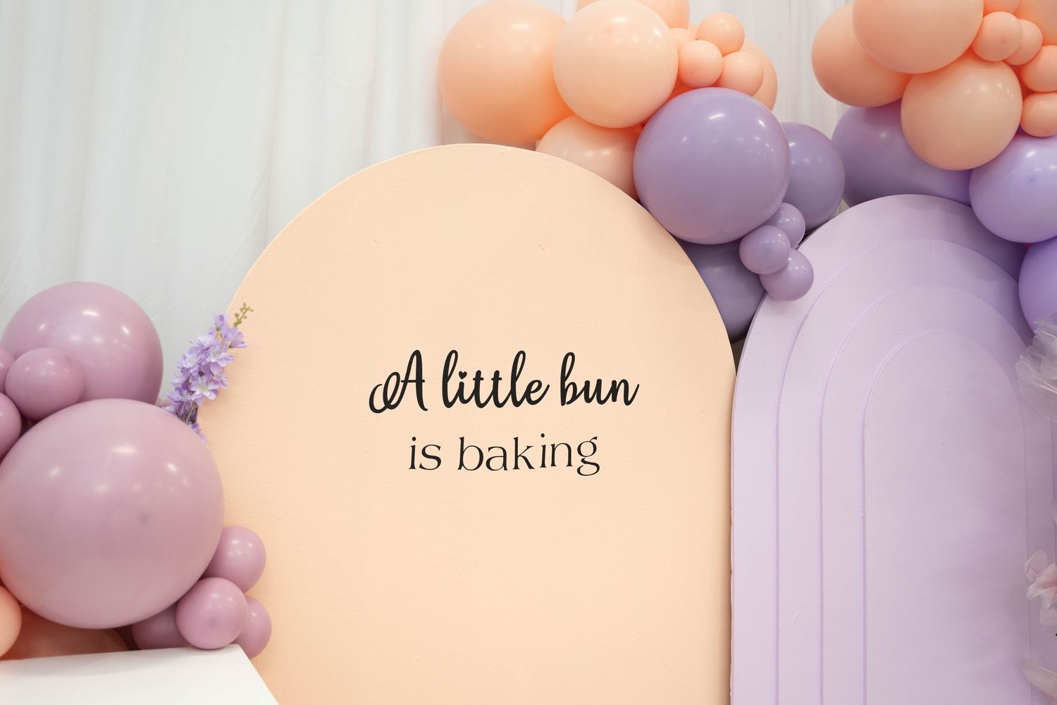Pastel peach and lavender balloon arch with a backdrop reading A little bun is baking for a baby shower.
