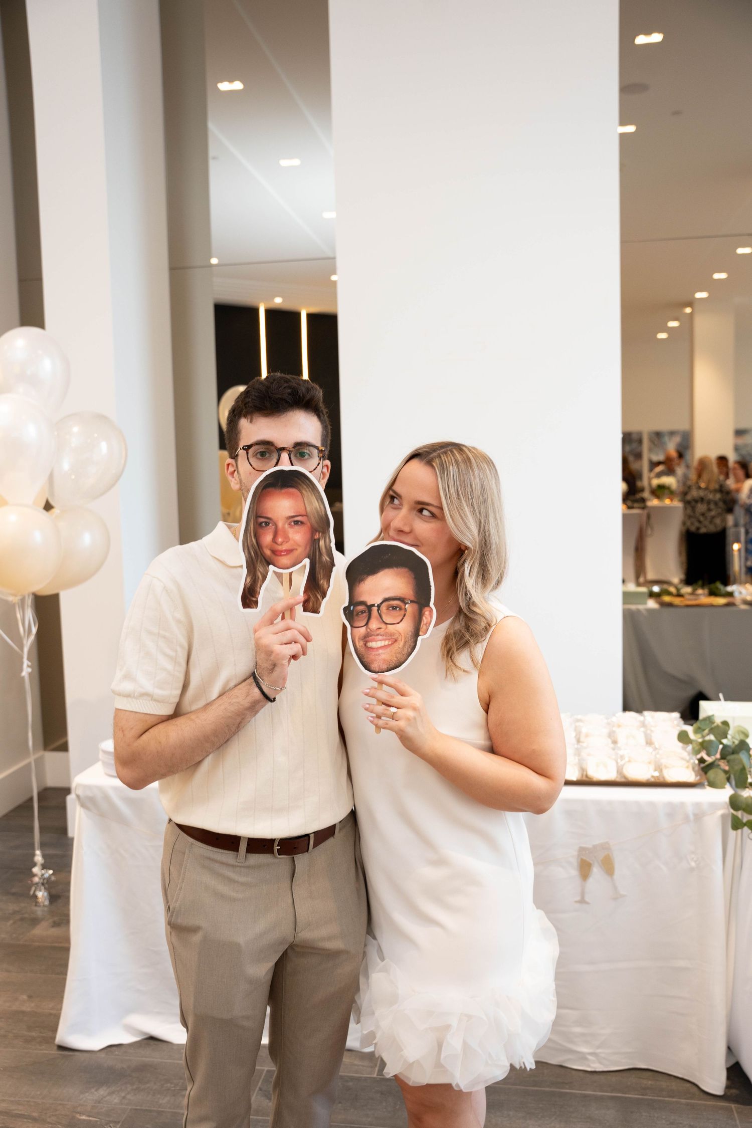 A smiling couple at an engagement party holds up cutout photo faces in front of their own faces beside balloons and a dessert table.