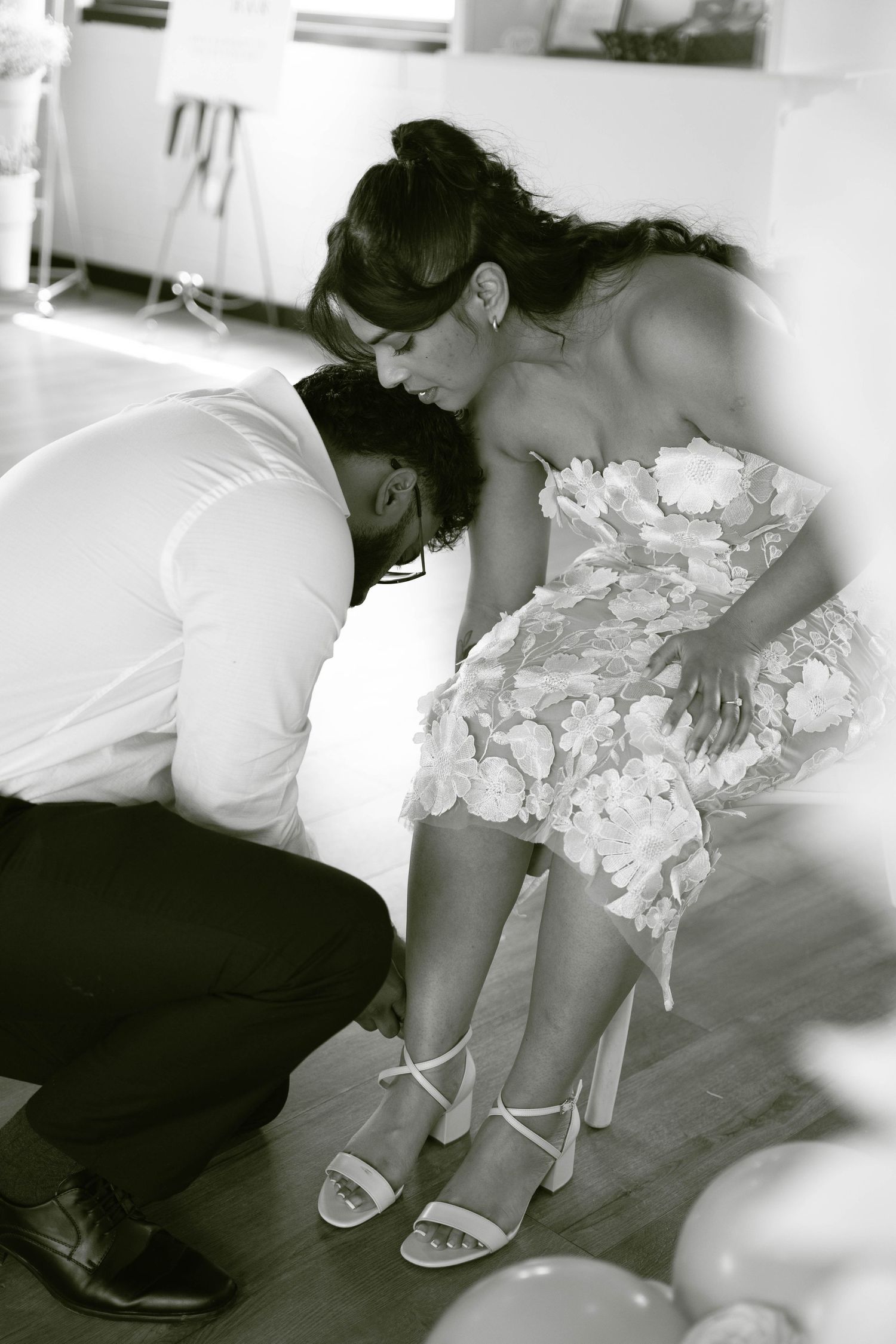 Black and white photo of a groom-to-be kneeling to buckle his fiancée's strappy sandals as she wears a floral lace dress.