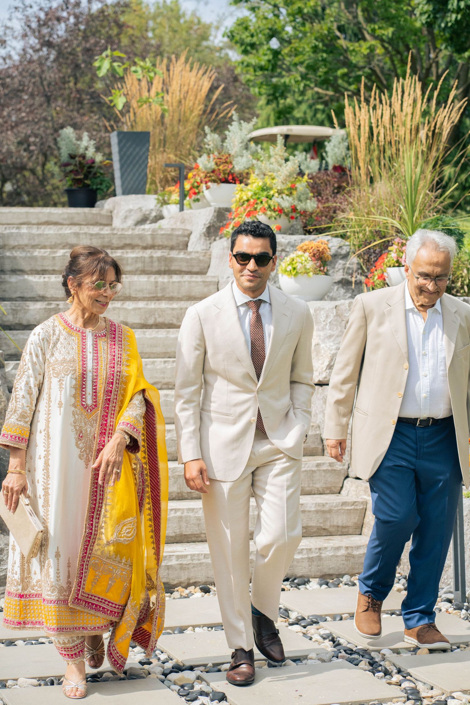 A groom in a beige suit and sunglasses walks down garden steps flanked by a woman in a yellow-trimmed outfit and an older man.
