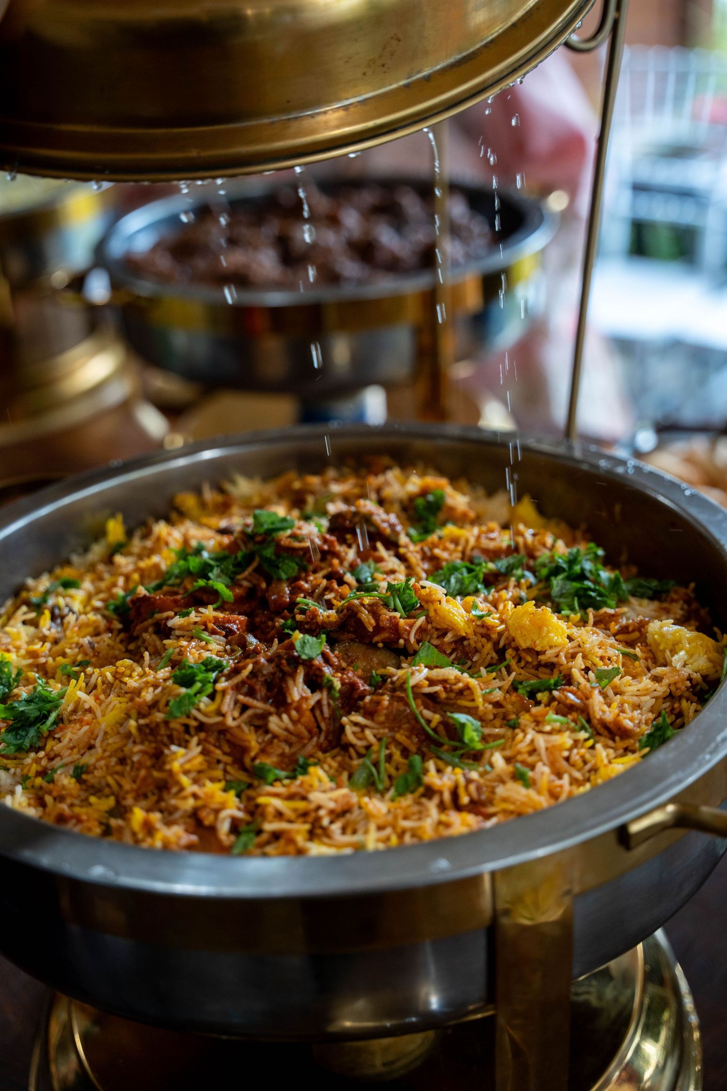 Steaming chafing dish filled with fragrant biryani garnished with fresh herbs and golden potatoes at a catered celebration.