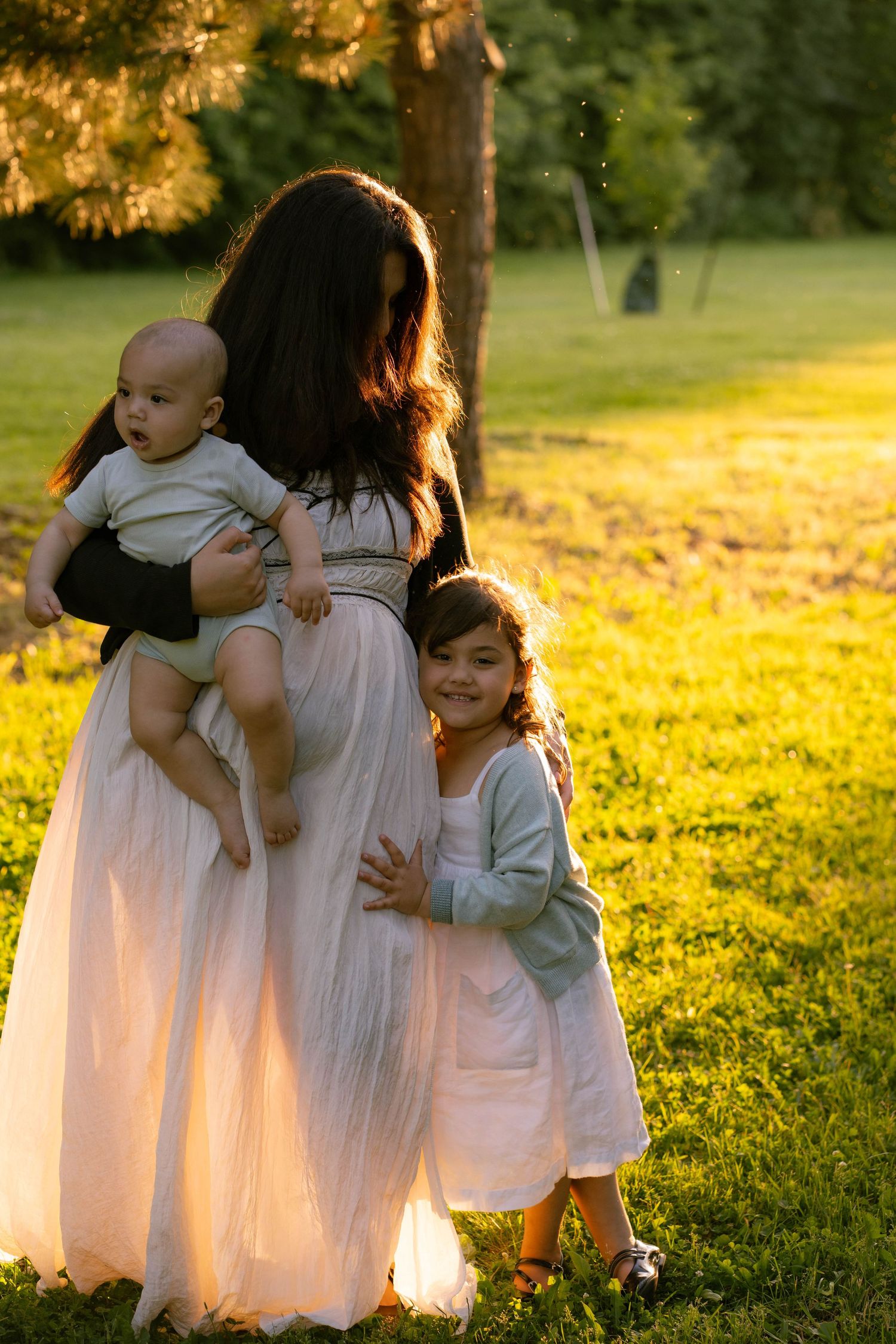 A mother in a flowing white dress holds a baby while a smiling little girl hugs her in a sunlit grassy park.