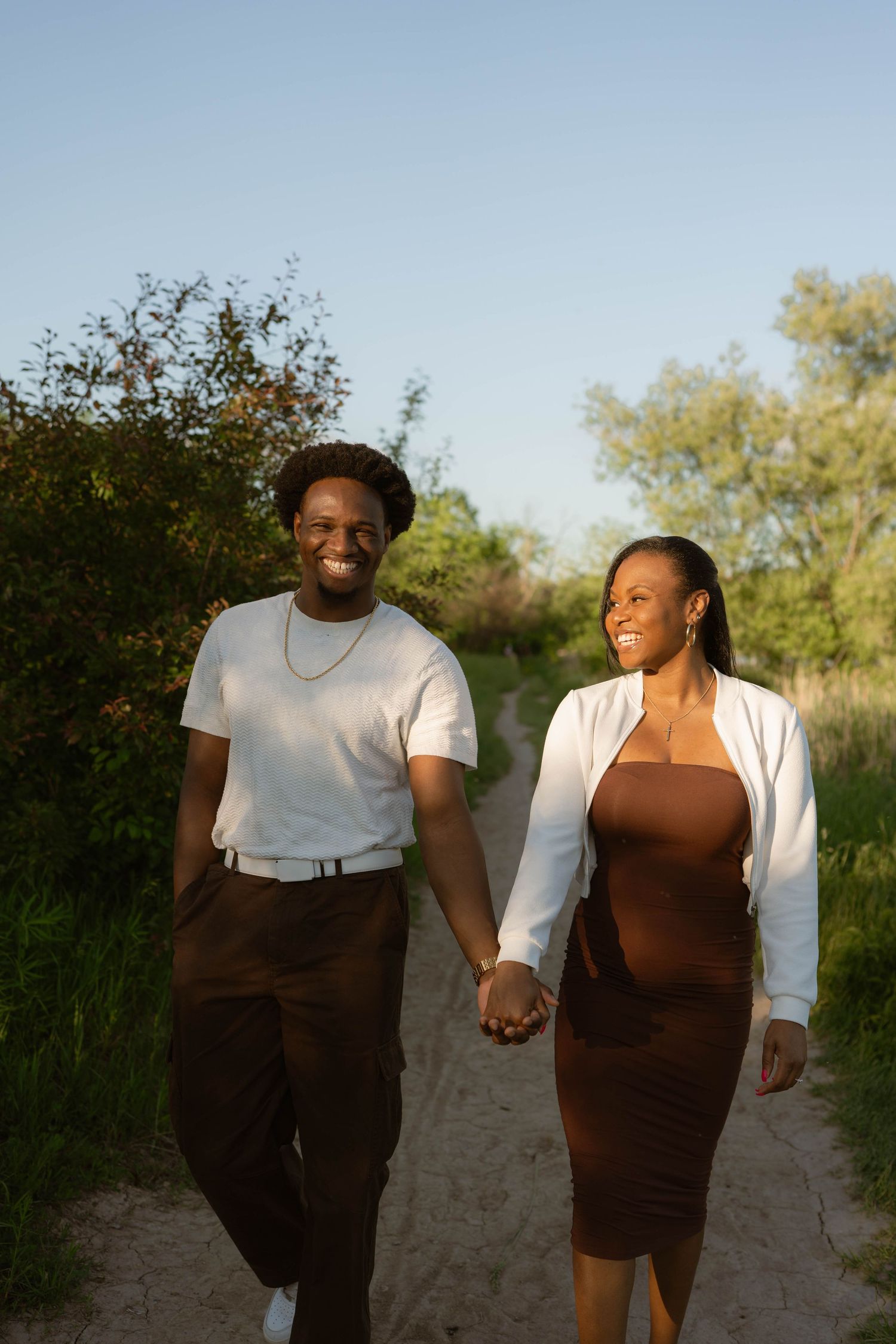 Smiling couple holding hands and walking along a sandy path surrounded by green trees in warm golden sunlight.