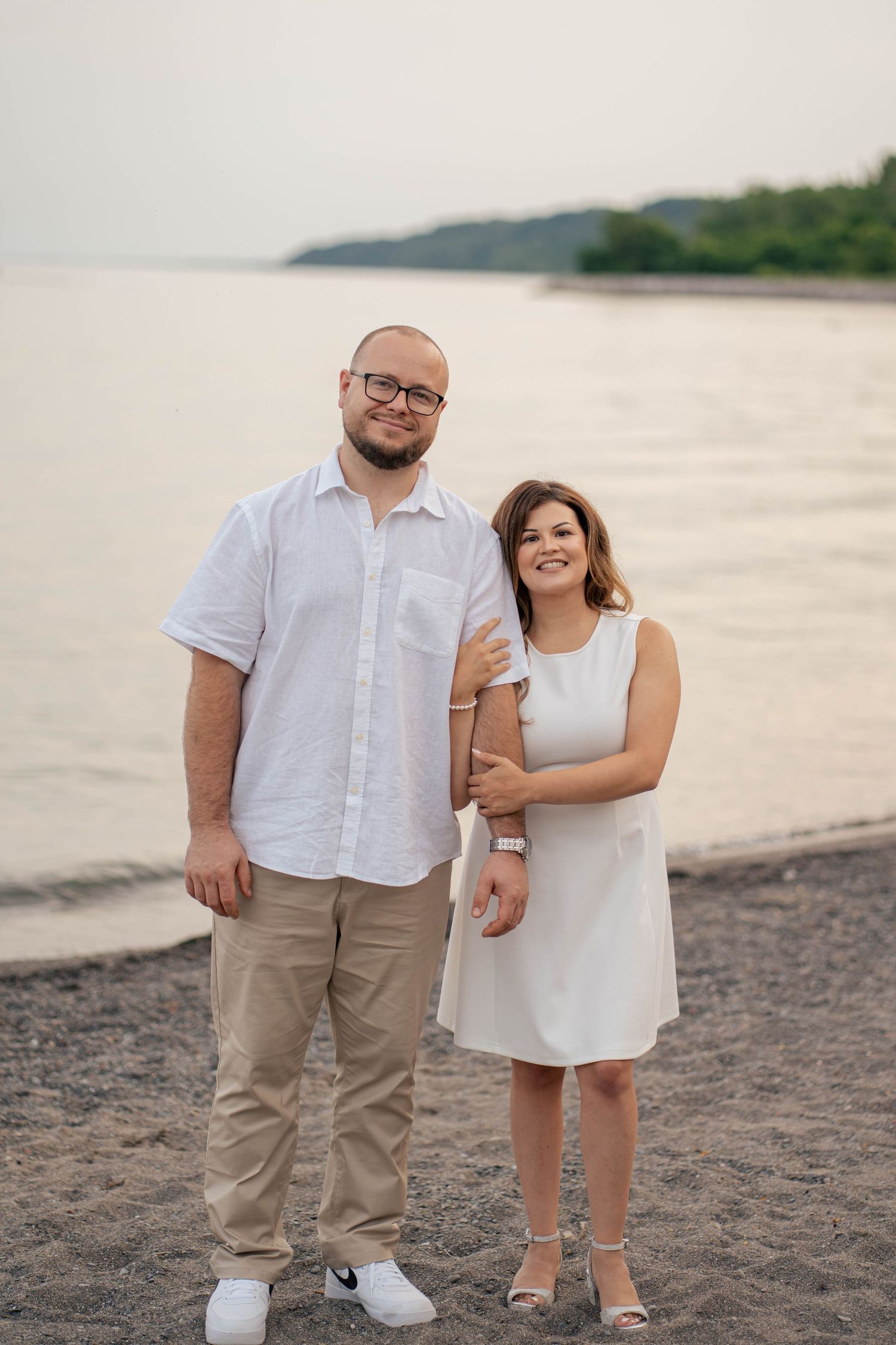 Smiling engaged couple in white standing arm in arm on a sandy beach with calm lake water and distant shoreline.