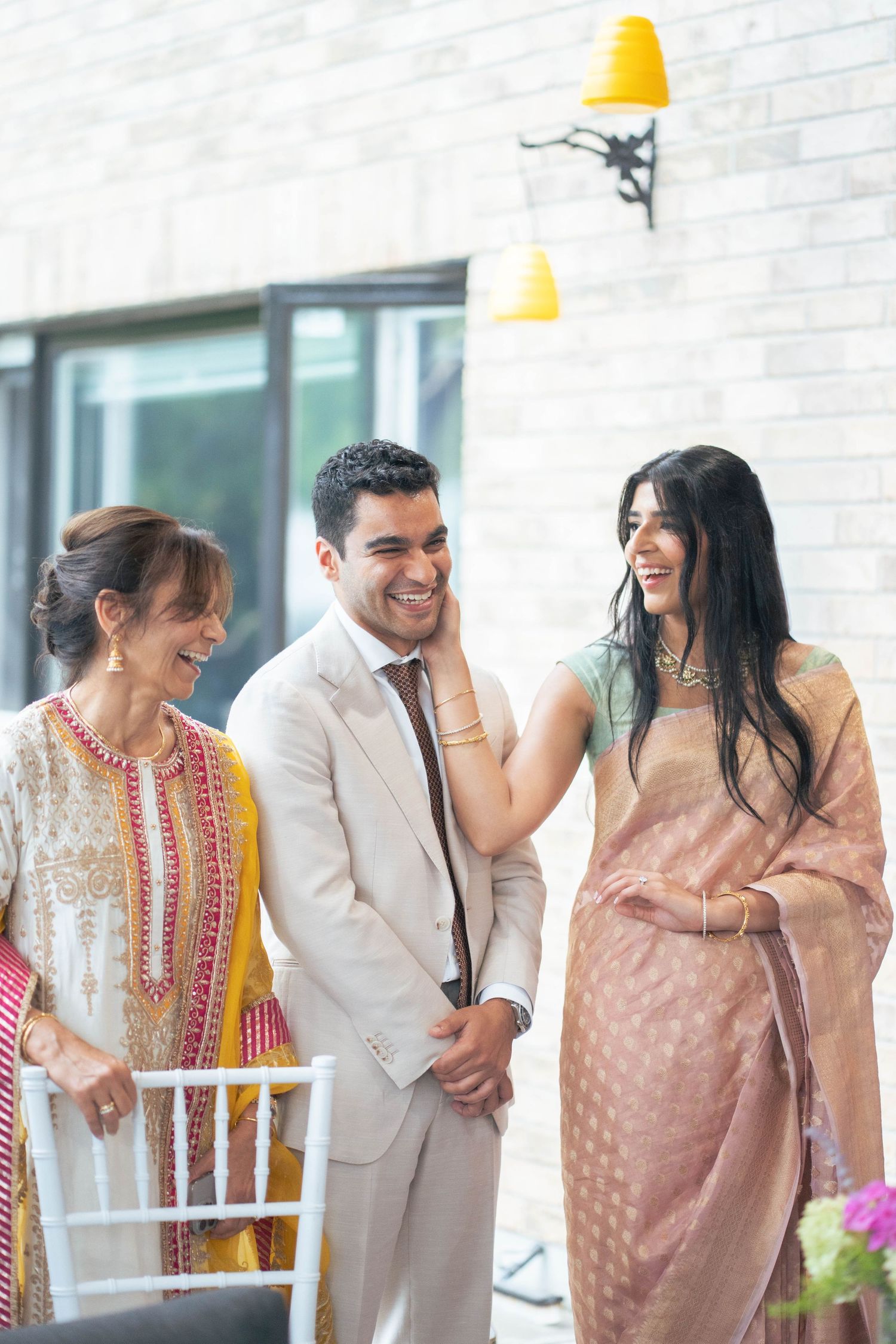 A laughing groom in a beige suit is lovingly touched on the cheek by his bride as an older woman smiles beside them.
