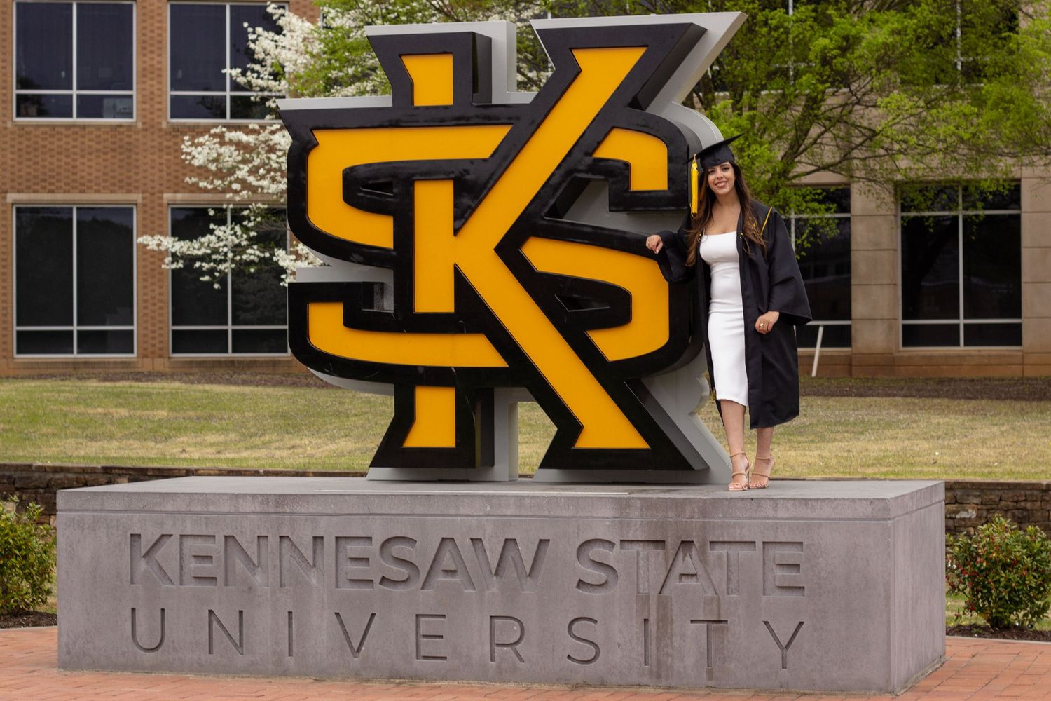Female graduate posing beside Kennesaw State University campus sign in cap and gown.